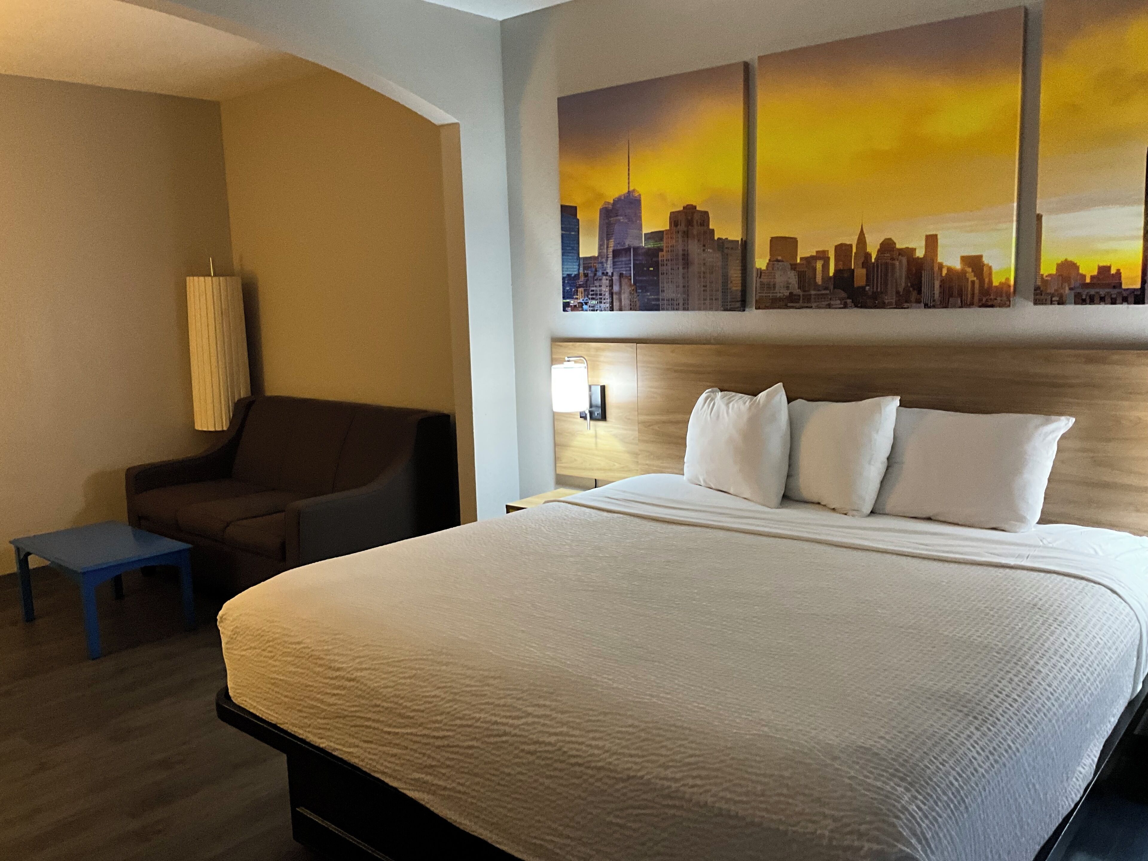 Une chambre d’hôtel avec un grand lit, une chaise, une petite table et un tableau représentant un paysage urbain accroché au mur.