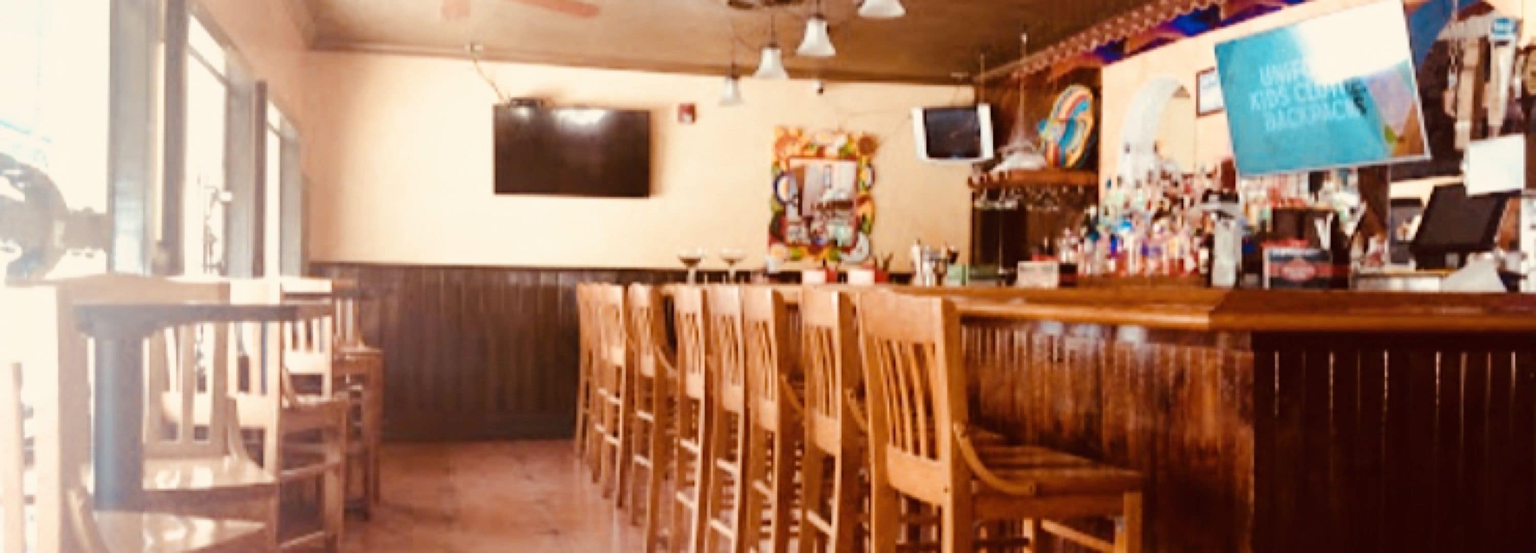 A bar area with wooden chairs and tables, a television mounted on the wall, and various bottles and glasses on the bar counter.