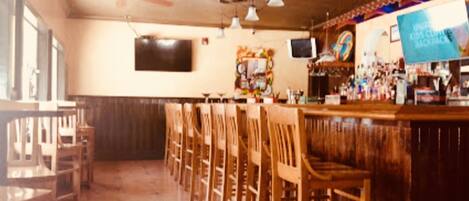 A bar area with wooden chairs and tables, a television mounted on the wall, and various bottles and glasses on the bar counter.