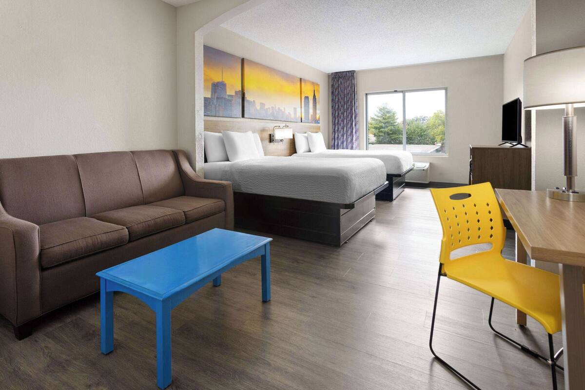 Habitación de hotel con una cama, un sofá, una mesa azul, una silla amarilla y un cuadro de un paisaje urbano en la pared.