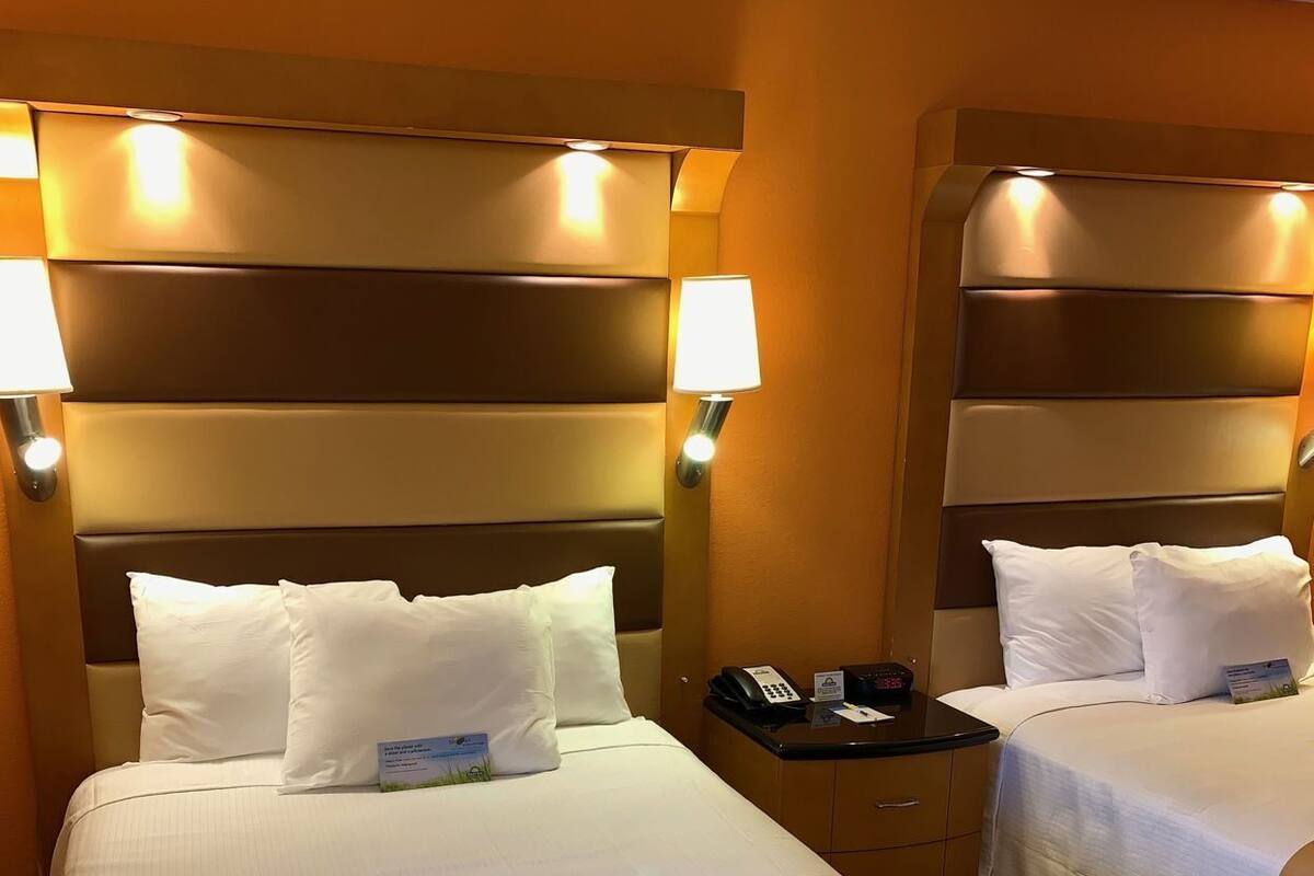 Habitación de hotel con dos camas, cada una con cabecera, lámparas de noche y un letrero que dice 'Hello Sunshine' en la colcha.
