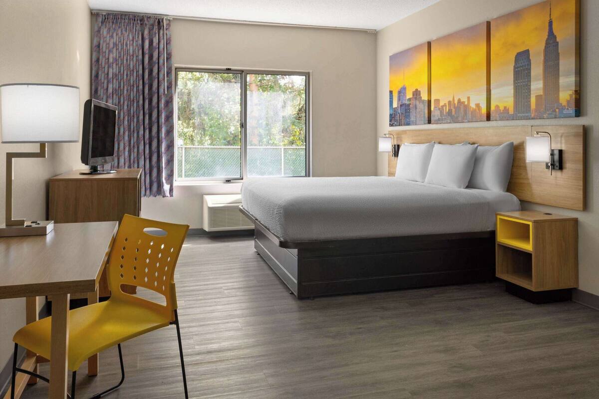 Una habitación de hotel con cama, escritorio, silla, televisión, ventana con cortinas y un cuadro con vista a la ciudad.