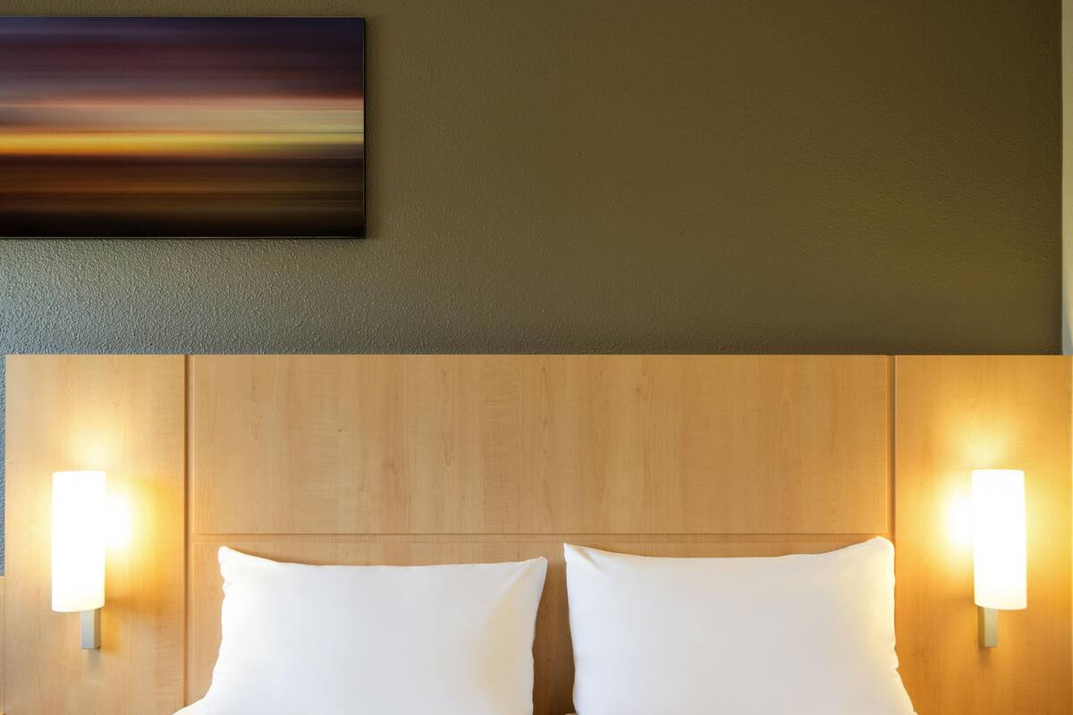 Hotellihuone, jossa on puinen pääty, kaksi valkoista tyynyä ja seinään kiinnitetyt valaisimet.