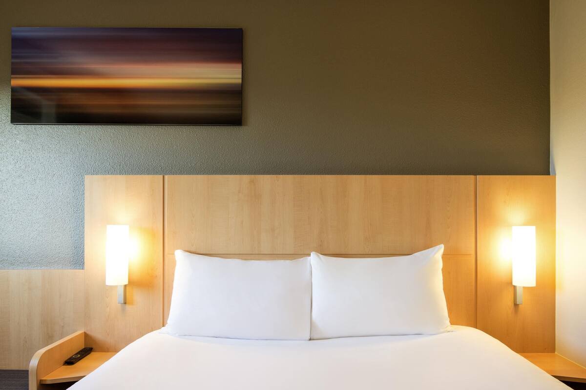 A hotel room with a wooden headboard, two bedside lamps, a white bed, and a painting on the wall.