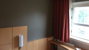 Huoneessa on puinen työpöytä, punaisilla verhoilla varustettu ikkuna ja seinälle asennettu käsidesipannin.