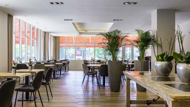 Un vaste espace intérieur aménagé en salle à manger, avec de nombreuses tables et chaises, de grandes fenêtres équipées de stores orange et des plantes en pot.