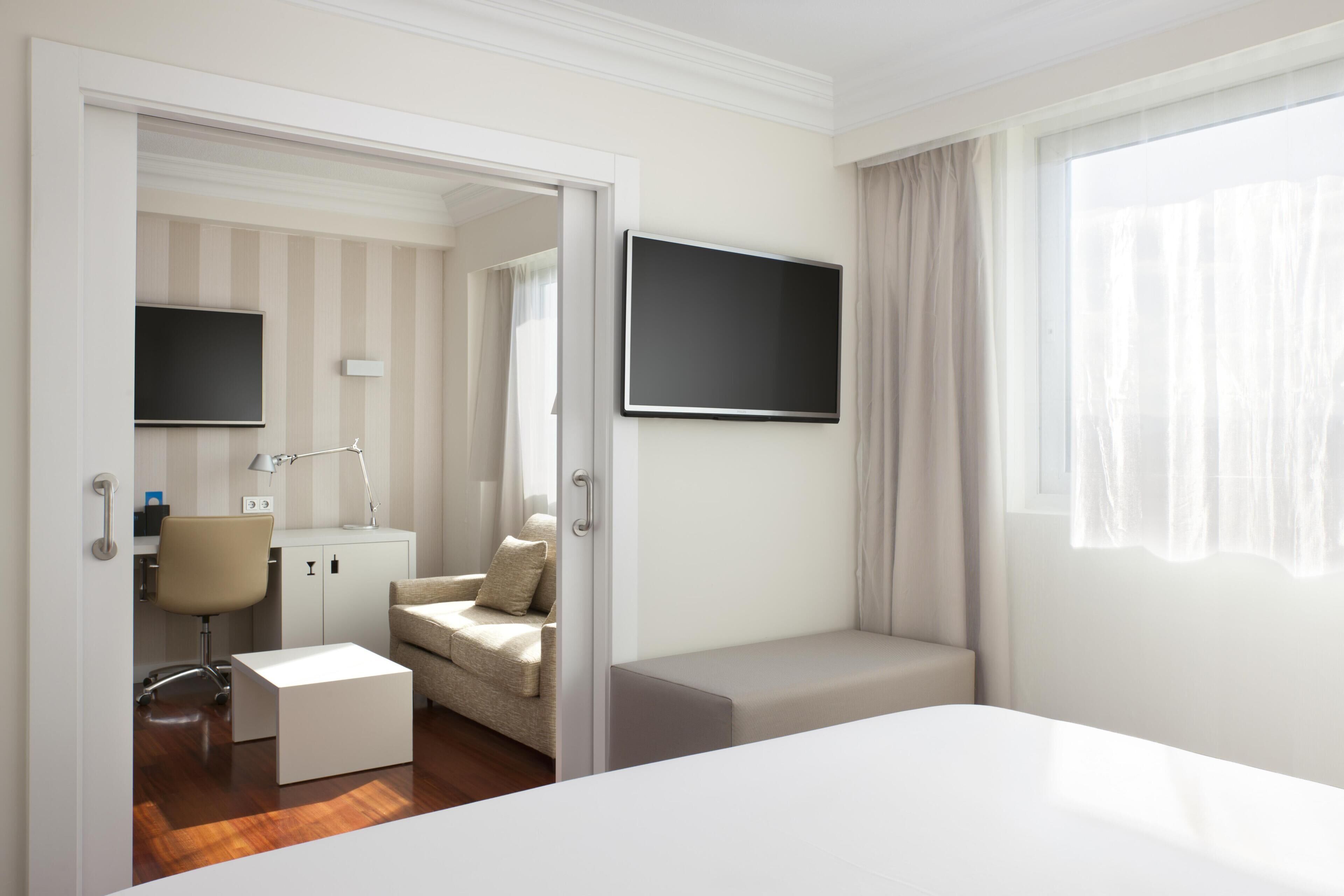 Ein Hotelzimmer mit einem Bett, einem Schreibtisch mit Stuhl, einem an der Wand montierten Fernseher und einem Fenster mit Vorhängen.