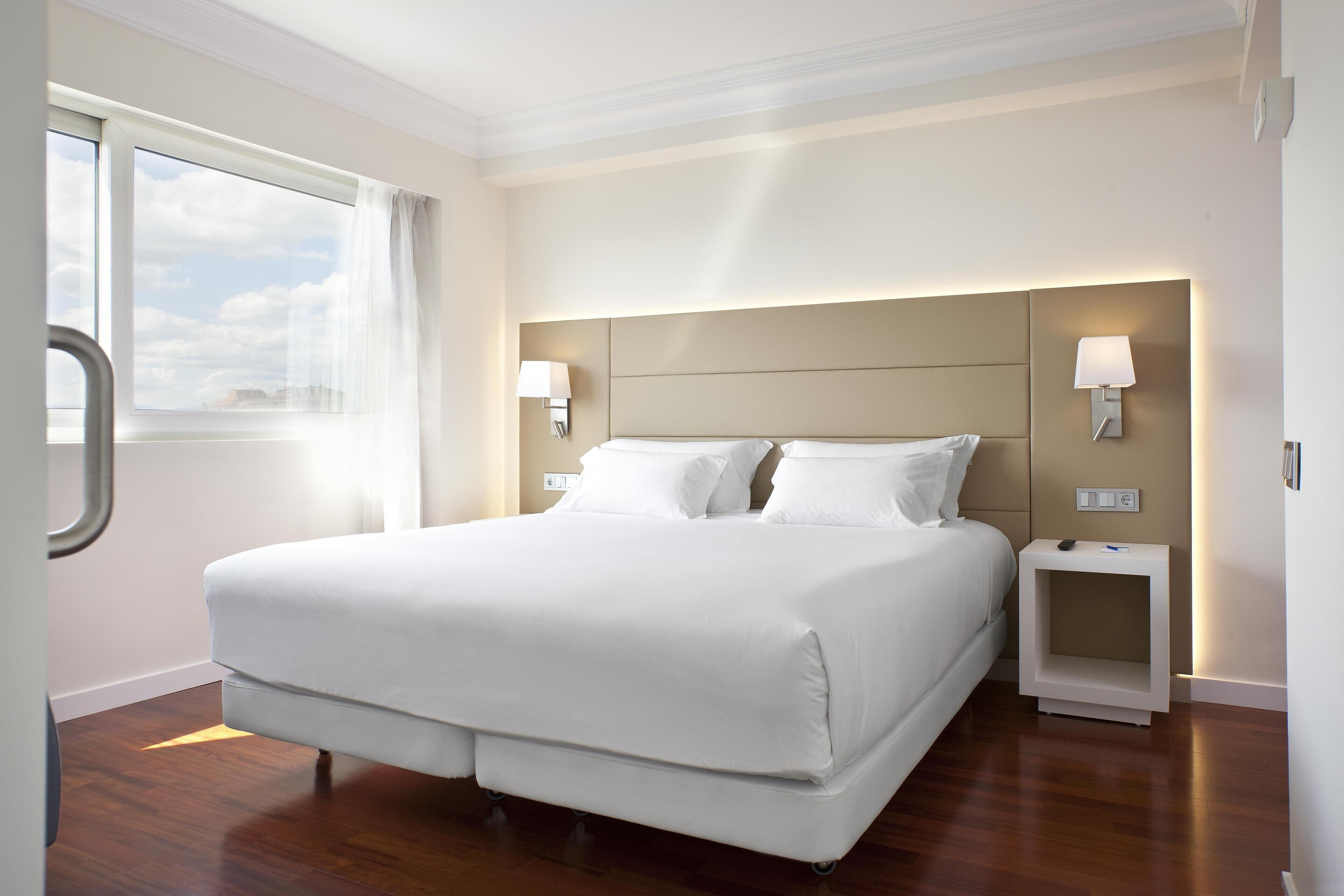 Una habitación de hotel moderna con una cama grande, dos mesitas de noche con lámparas, una ventana con cortinas y suelo de madera.