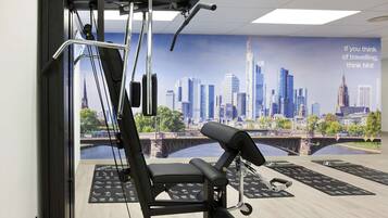 Un gimnasio con equipamiento de ejercicio, un mural de paisaje urbano y colchonetas en el suelo.