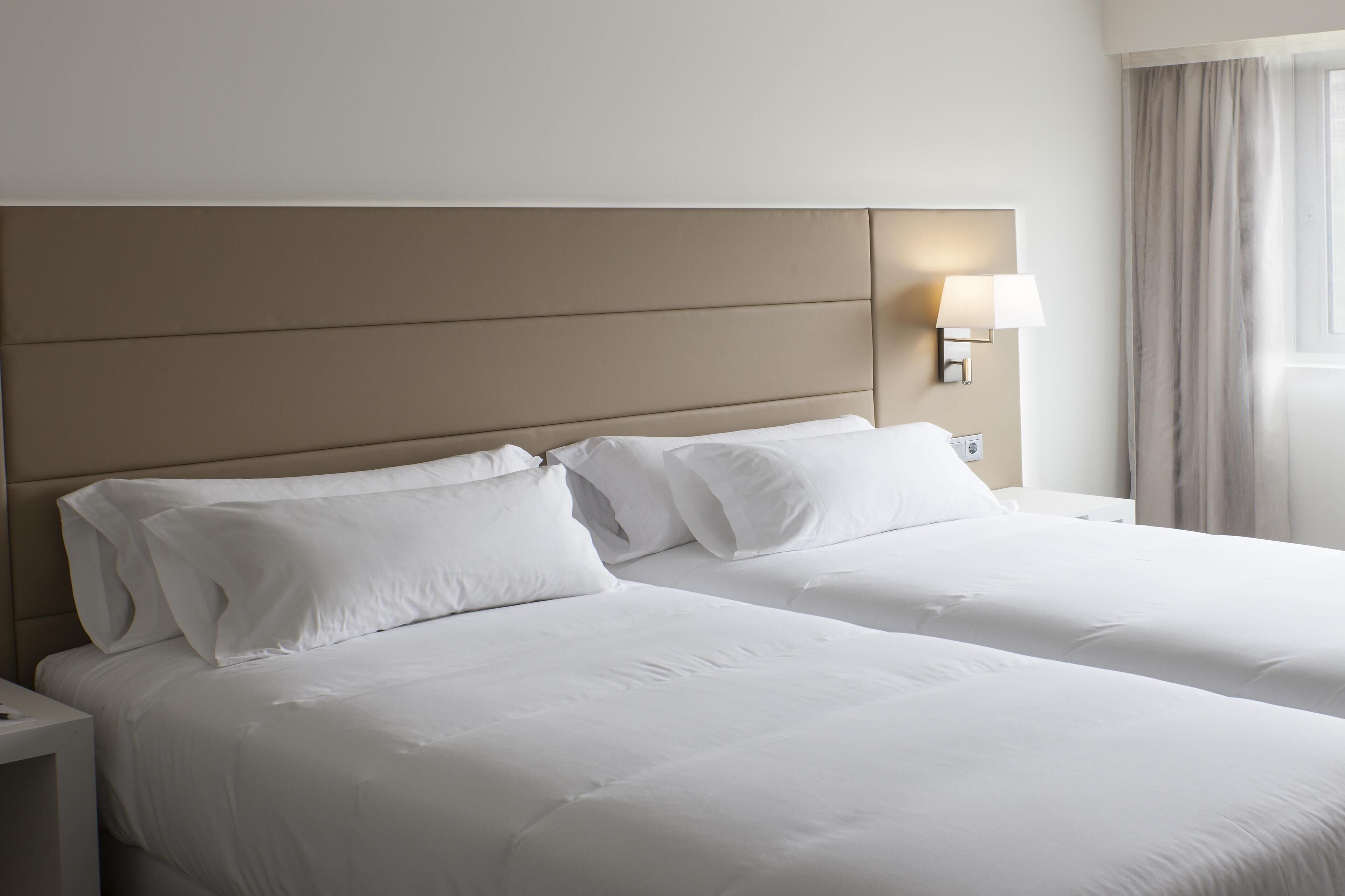 A hotel room with two beds, a nightstand, a lamp, and a window with curtains.