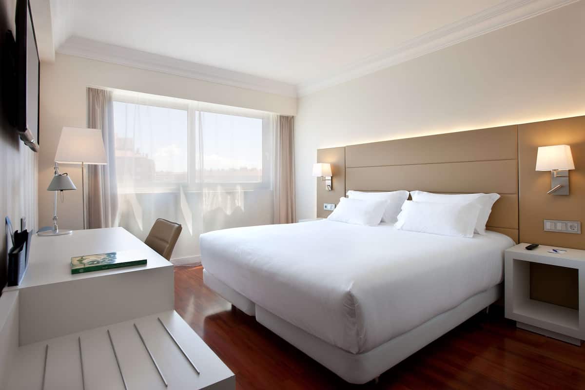 Une chambre d’hôtel avec un grand lit, des tables de chevet, un bureau avec une lampe et une vue sur la ville par la fenêtre.