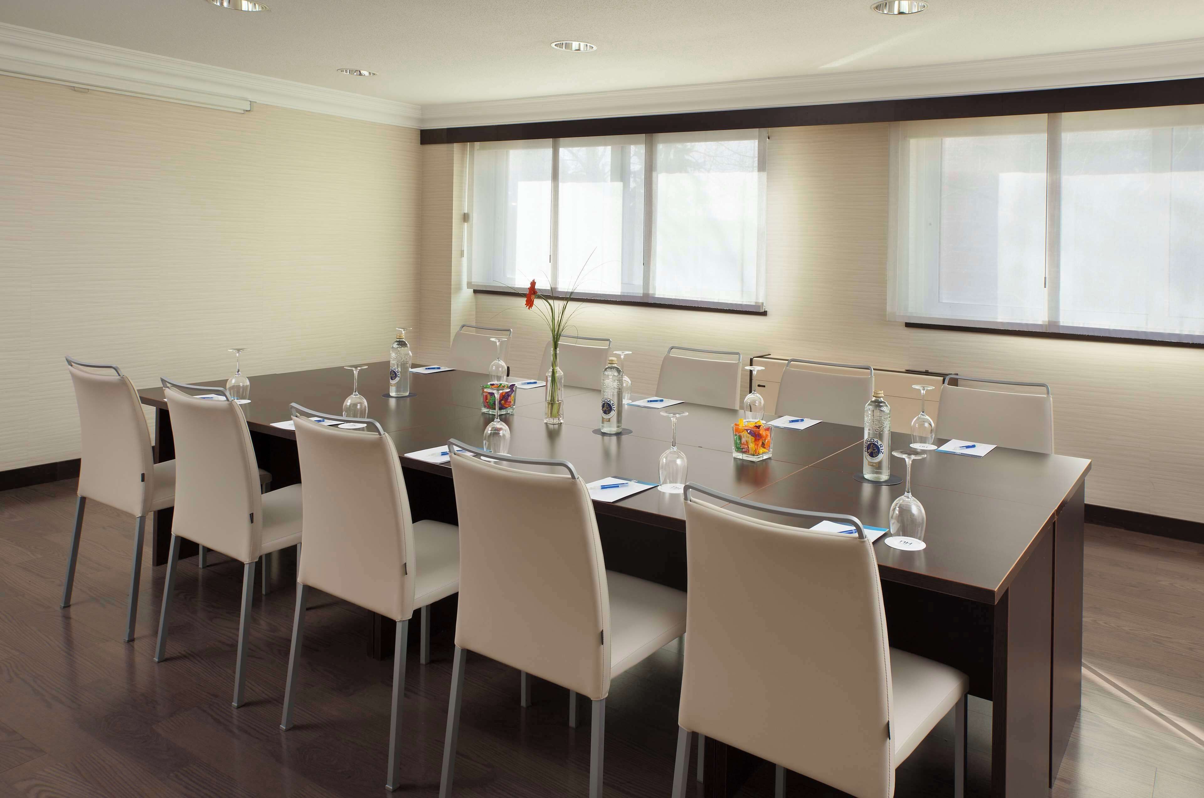 Un salón de conferencias con una mesa de madera oscura, sillas blancas y un jarrón con flores.