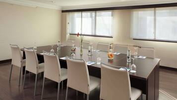 Un salón de conferencias con una mesa de madera oscura, sillas blancas y un jarrón con flores.