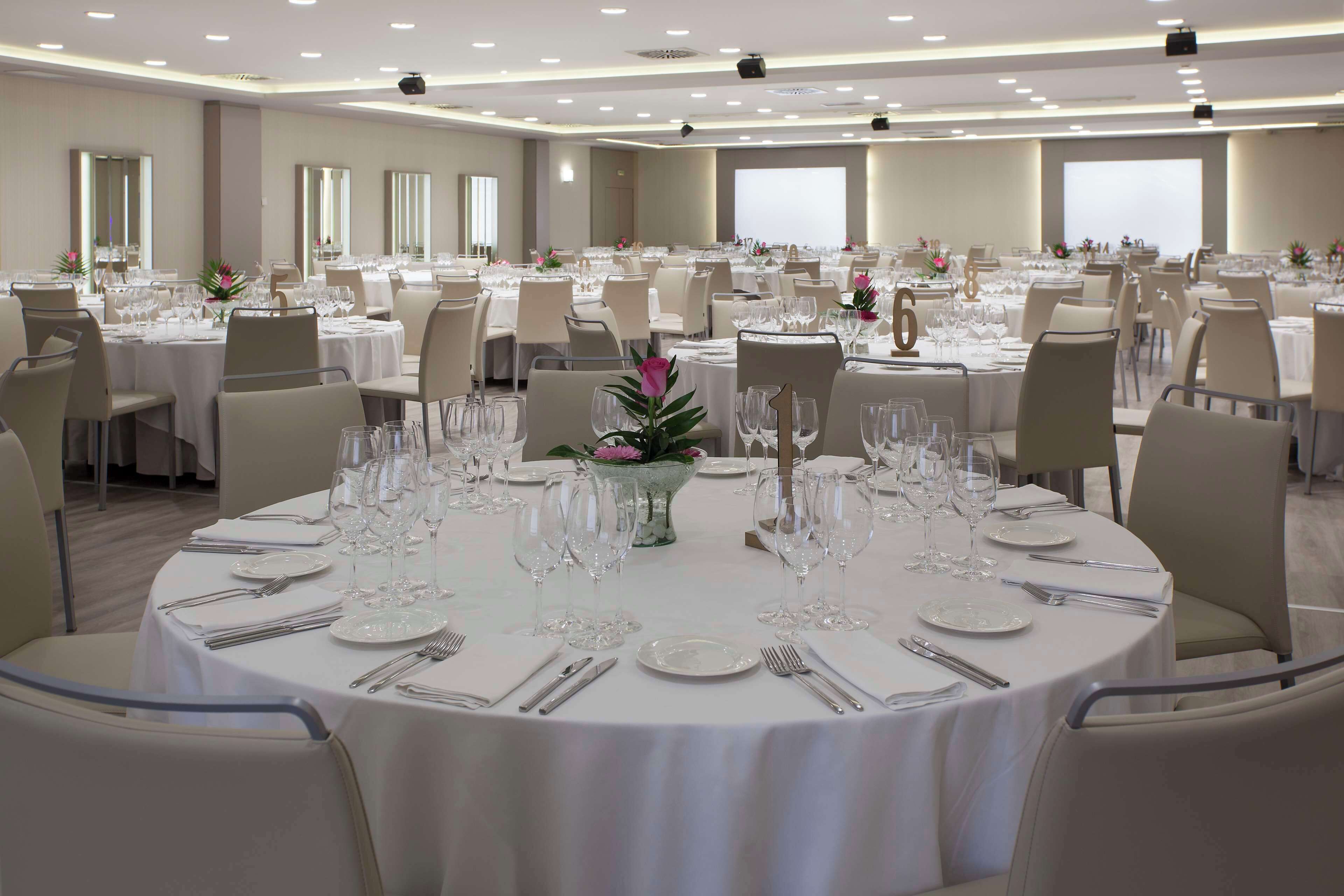 Un salón de banquetes con mesas redondas dispuestas para comer, cada una con platos, cubiertos y copas de vino.