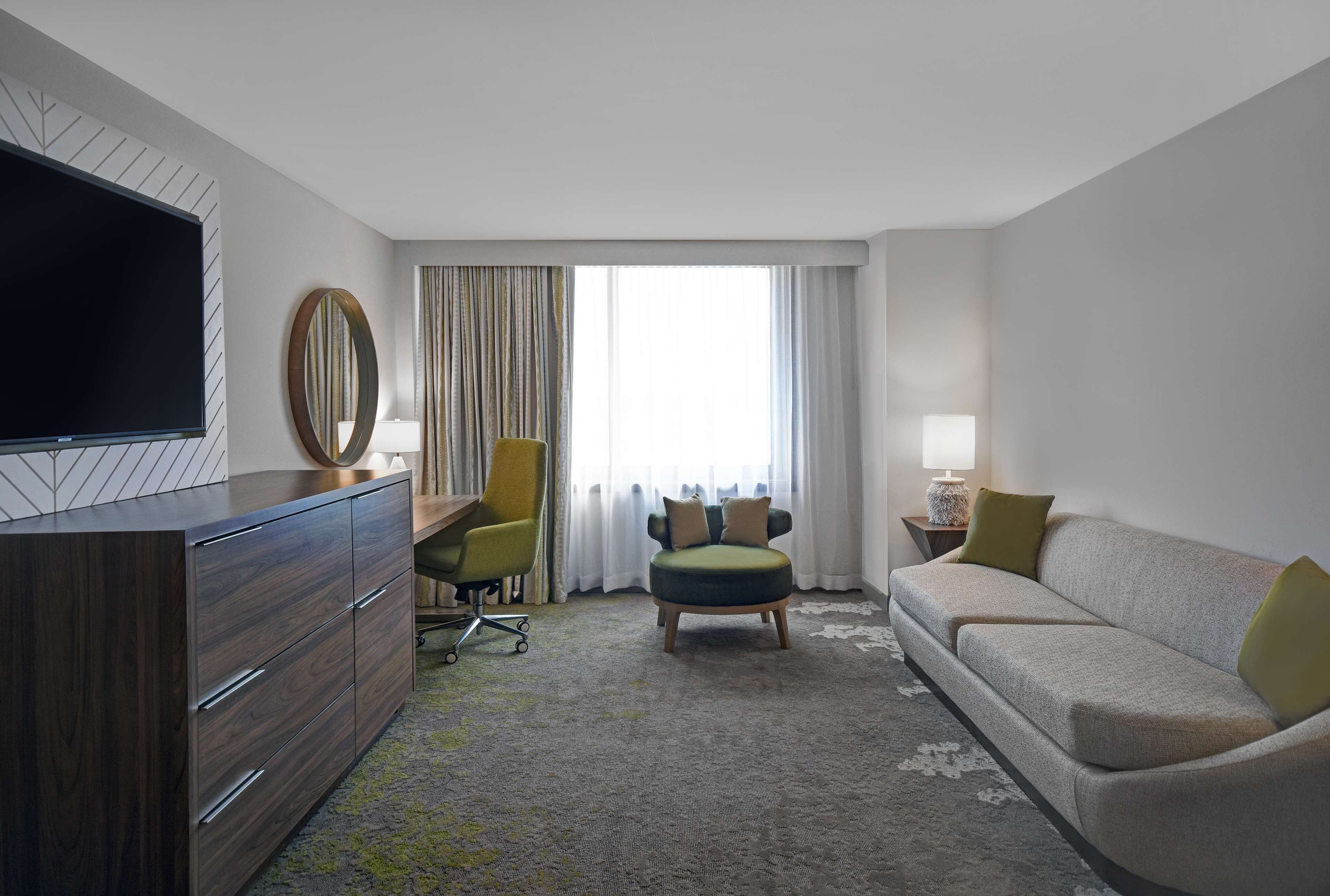 A hotel room with a flat-screen TV, a wooden dresser, a green armchair, a sofa, and a large window with curtains.