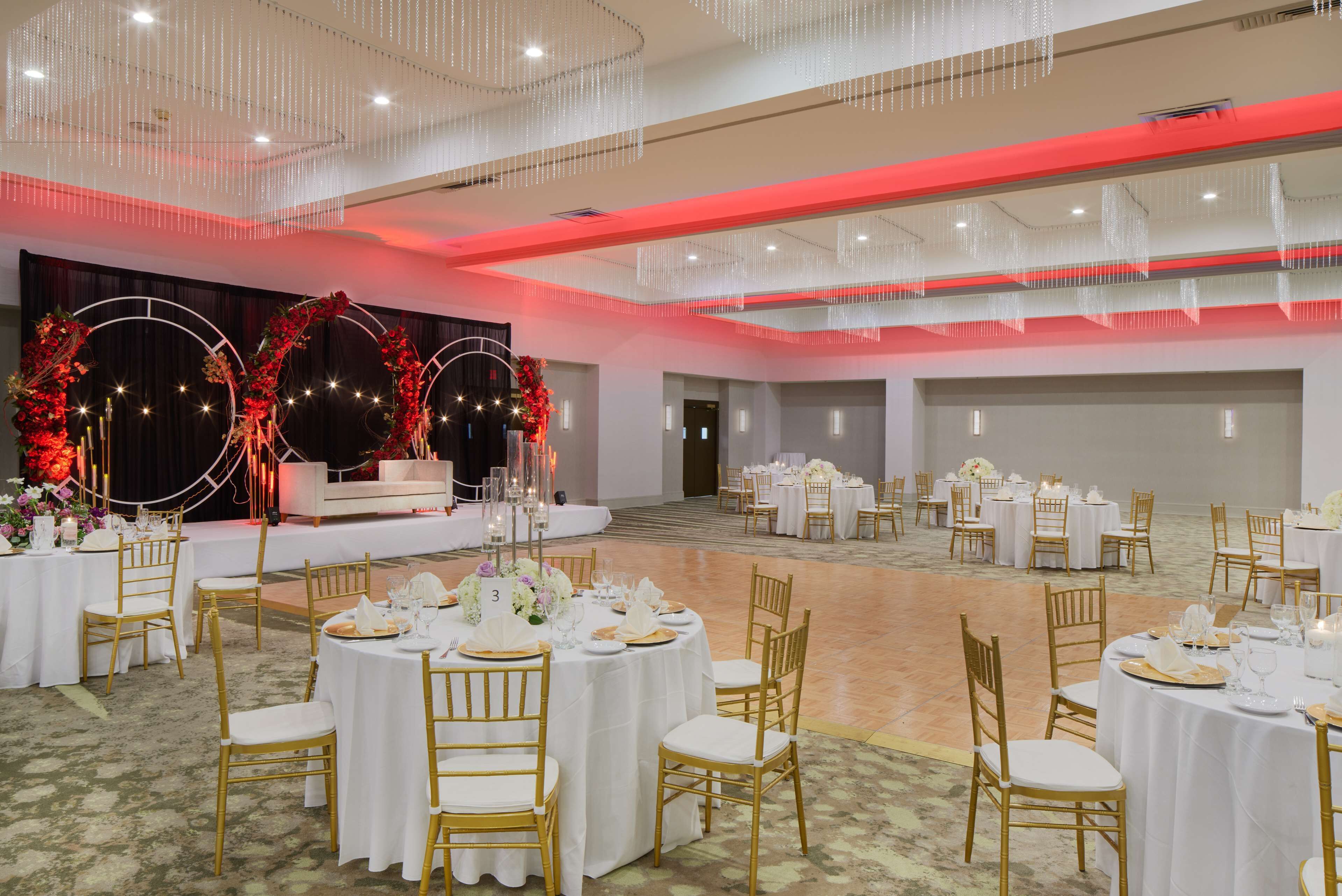 A banquet hall with round tables, gold chairs, and a stage with floral decorations.