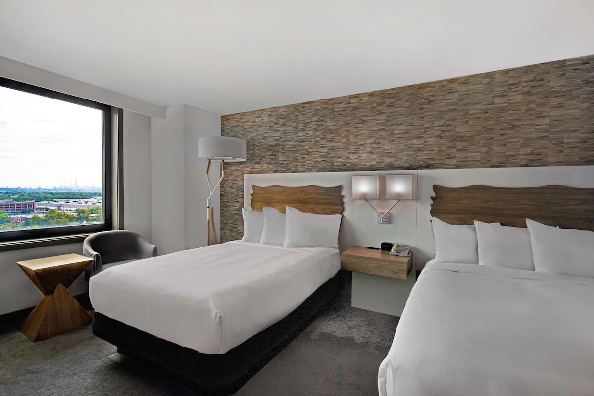 Een hotelkamer met een groot bed, een nachtkastje, een stoel en uitzicht op de stad door een groot raam.