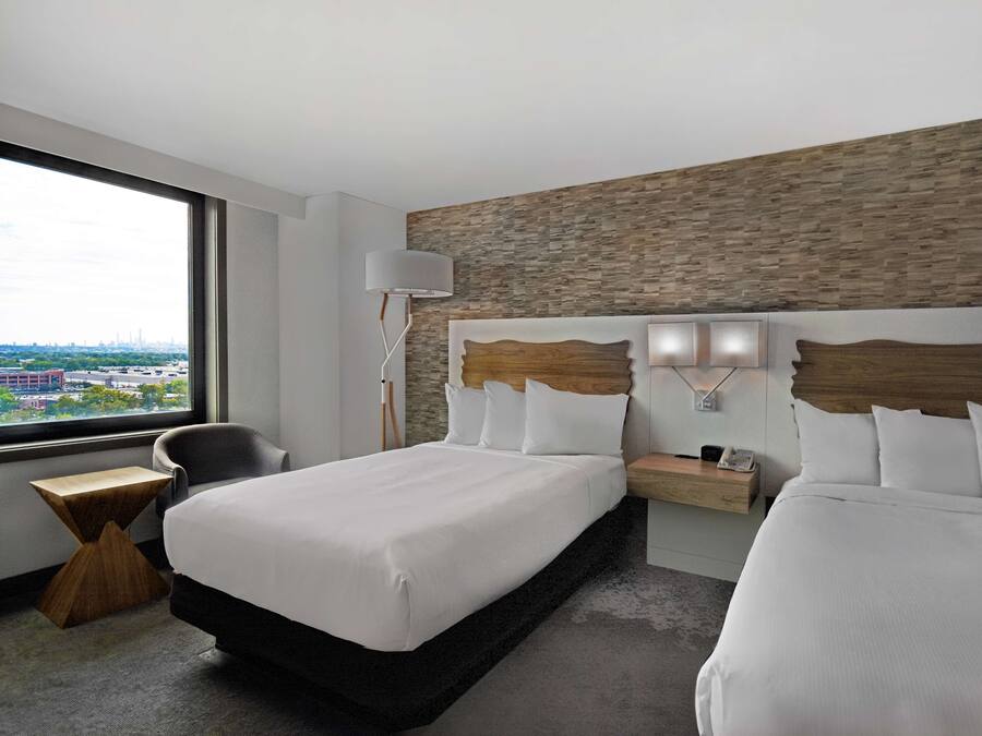 A hotel room with a large bed, a bedside table, a chair, and a view of the city through a large window.
