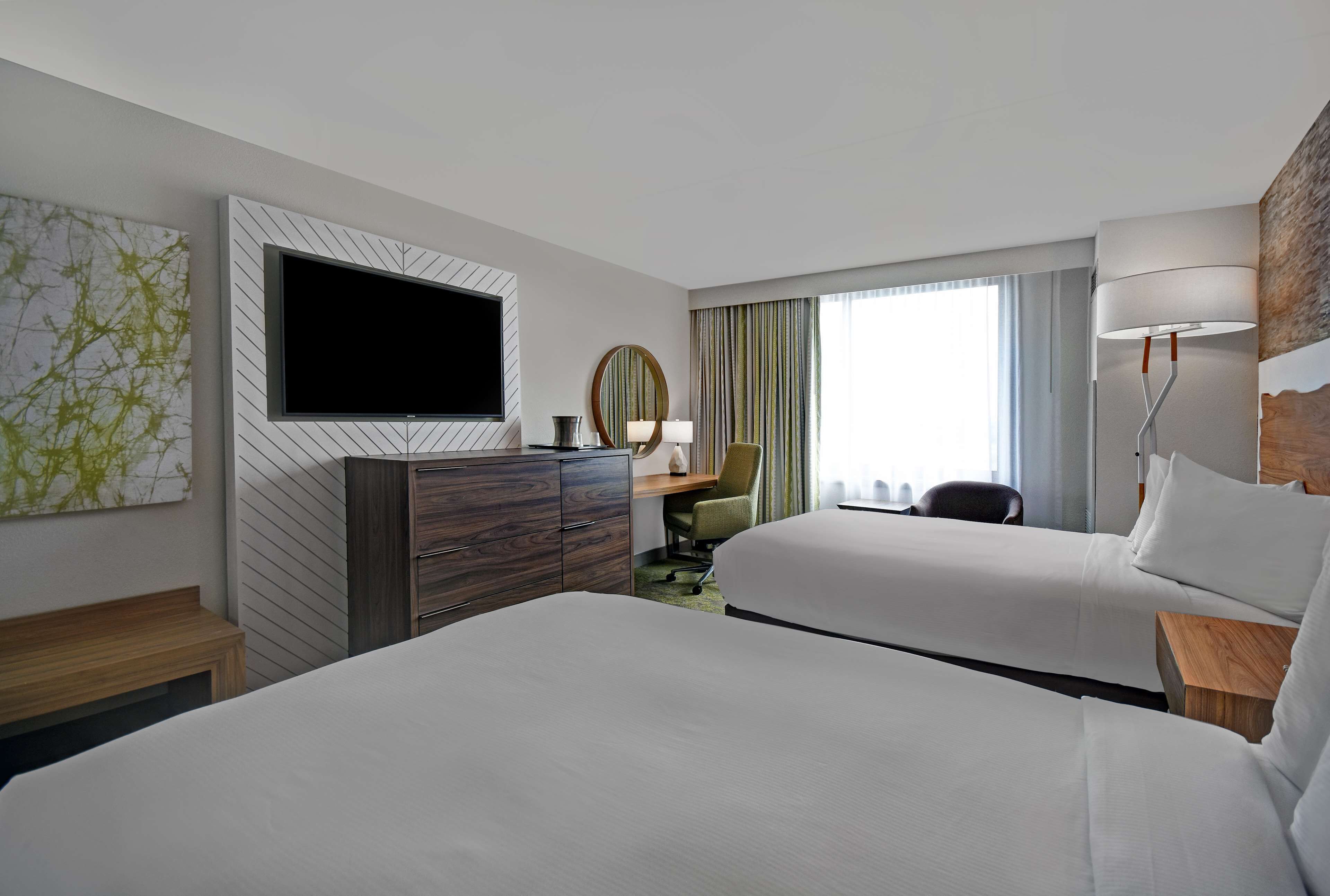 A hotel room with two beds, a flat-screen TV mounted on the wall, a desk with a chair, and a large window with curtains.