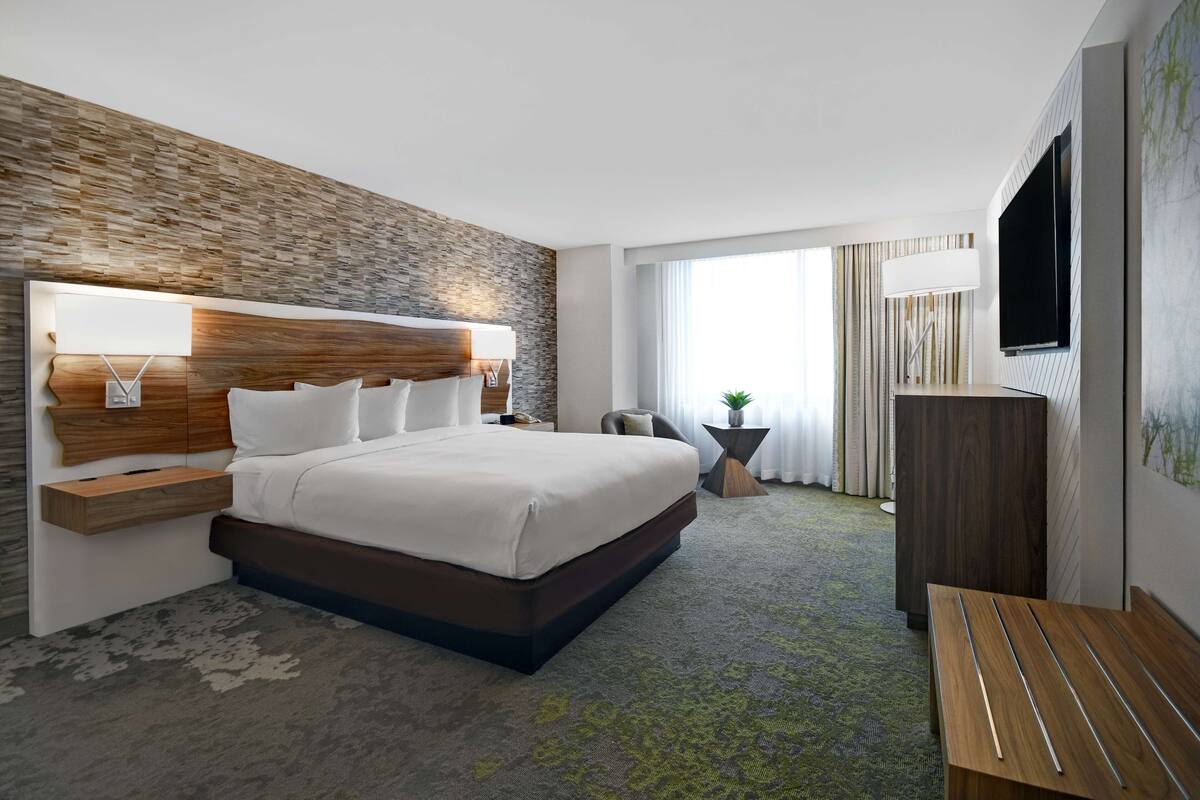 Een moderne hotelkamer met een groot bed, een houten hoofdeinde, een flatscreen televisie en een groot raam met uitzicht op de omgeving.
