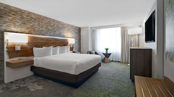 A modern hotel room with a large bed, a wooden headboard, a flat-screen TV, and a large window offering a view of the outdoors.