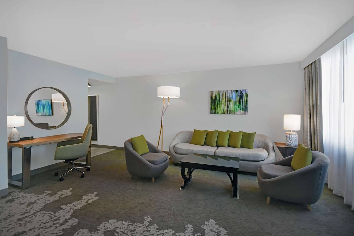 Een moderne hotelkamer met een bureau, twee fauteuils, een bank, een bijzettafeltje en een groot raam met gordijnen.