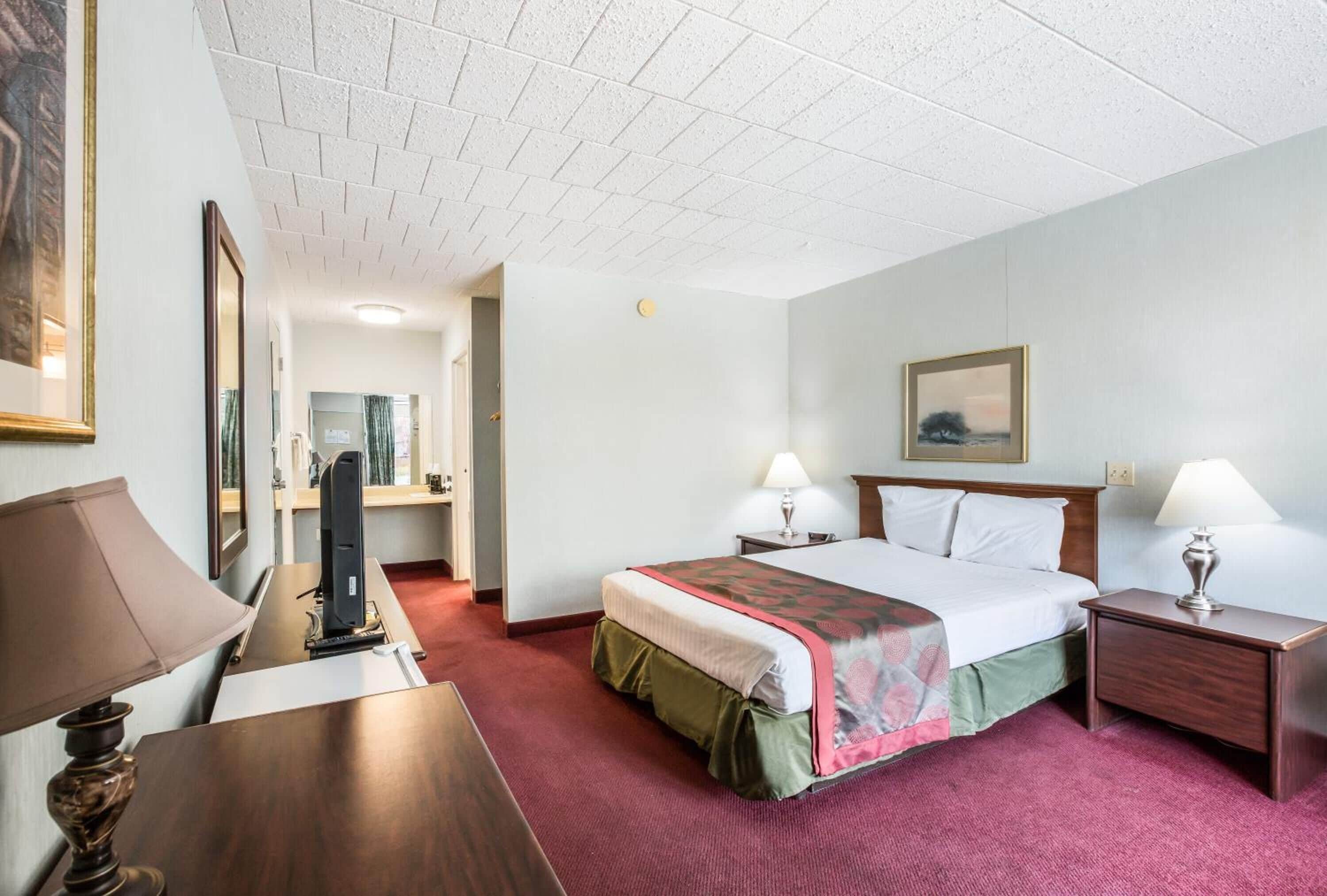 Habitación de hotel con cama, mesitas de noche, escritorio, lámpara, espejo y un cuadro en la pared.