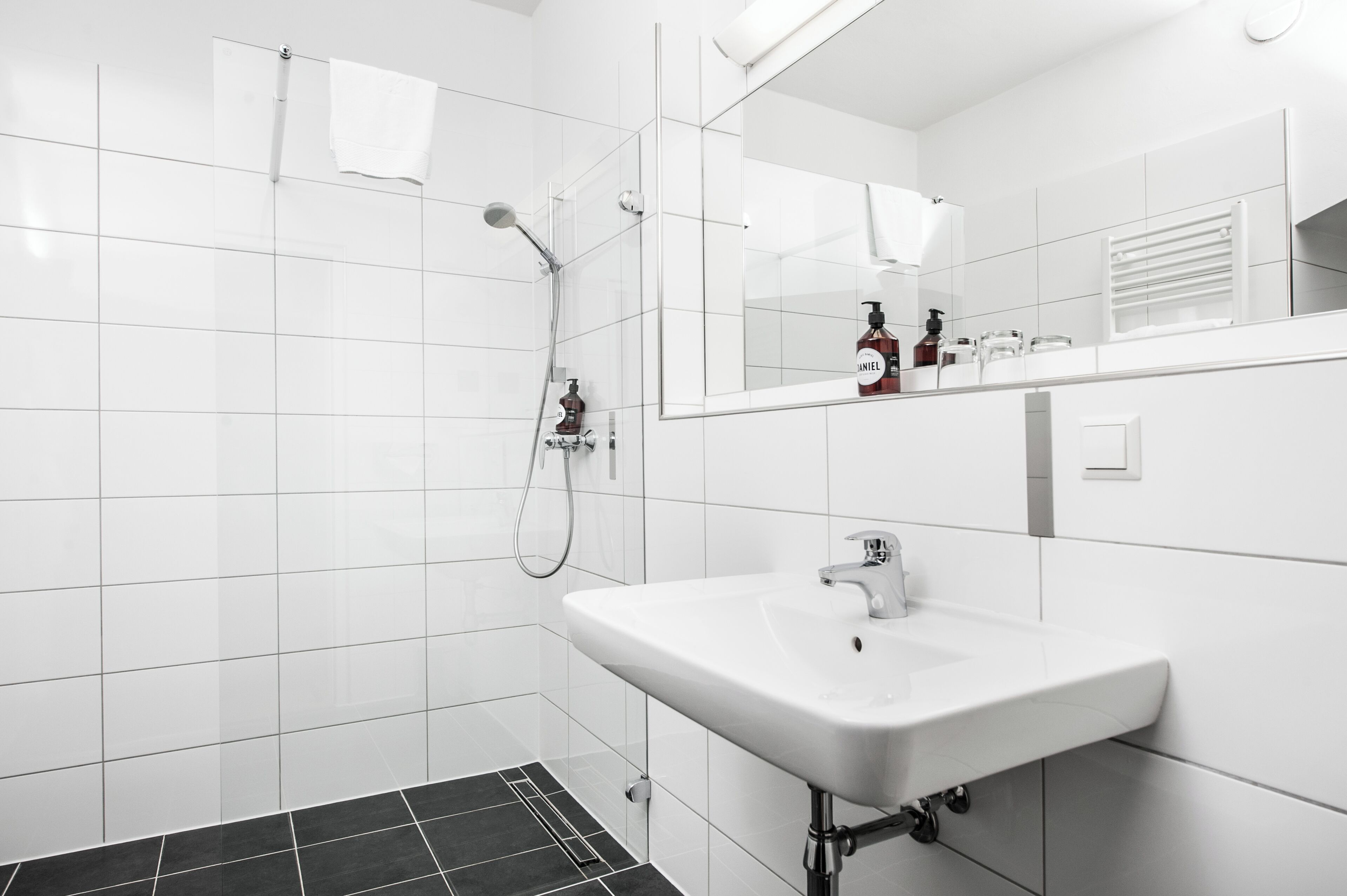 Un baño moderno con azulejos blancos, un lavamanos blanco, un espejo y una ducha.