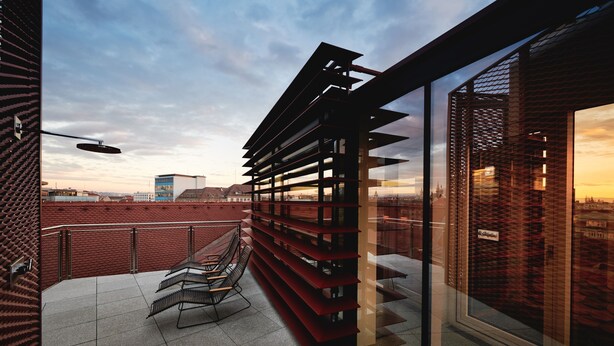 Un edificio moderno con fachada de ladrillo rojo, un balcón de vidrio con una banca de metal y vistas del horizonte urbano.