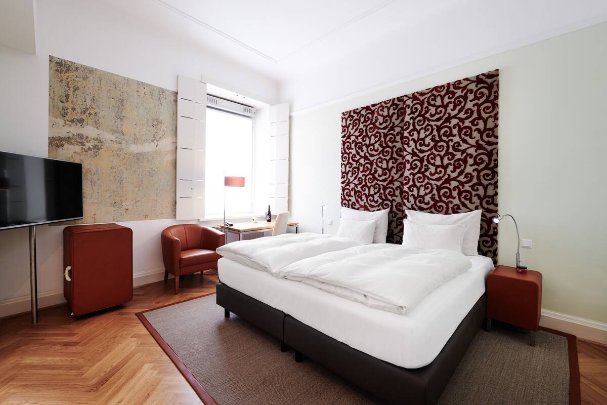 A modern hotel room with a large bed, a desk, a chair, a TV, and a patterned headboard.