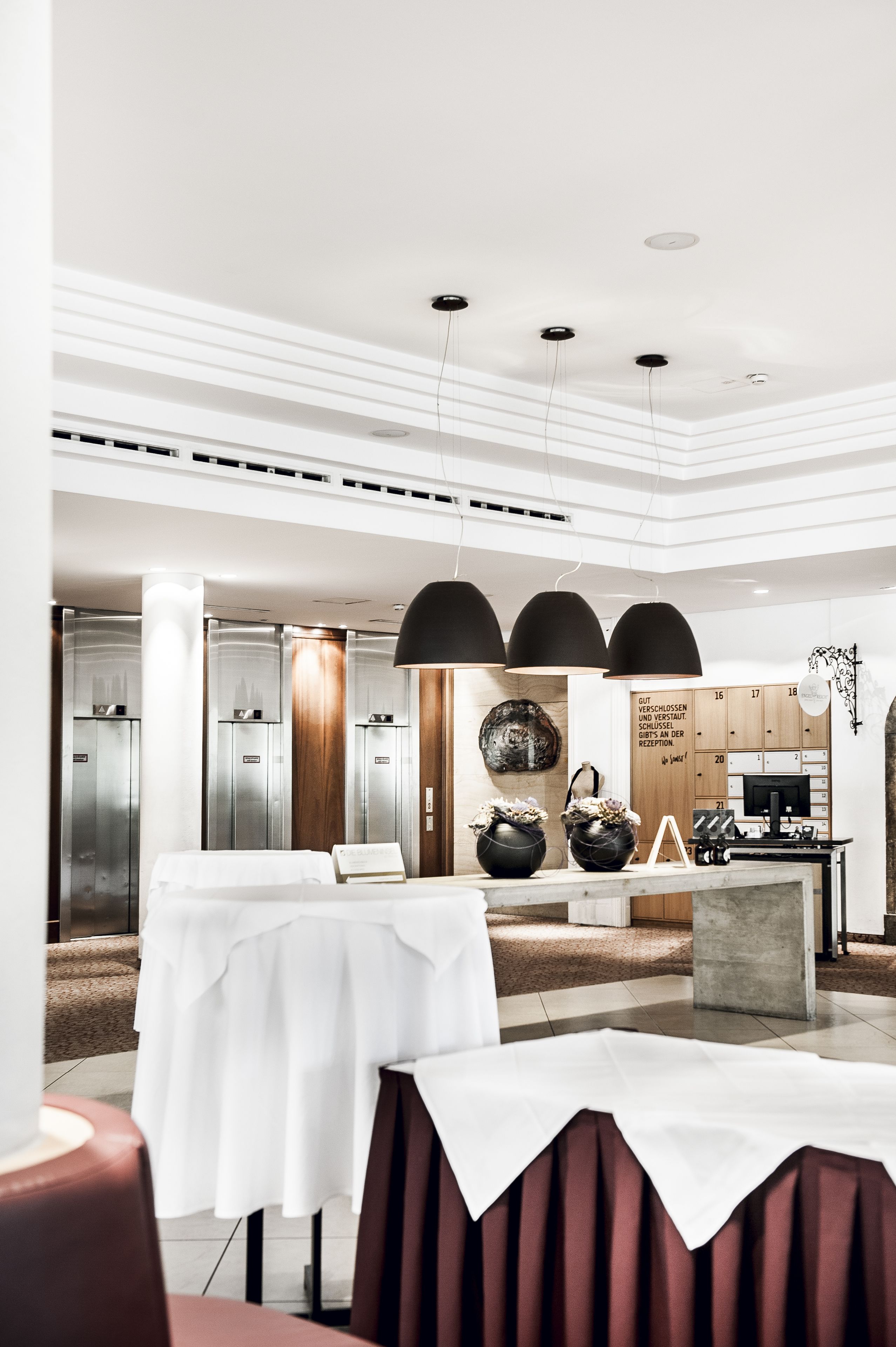 A modern hotel lobby with a reception desk, elevators, and a dining area with tables and chairs.