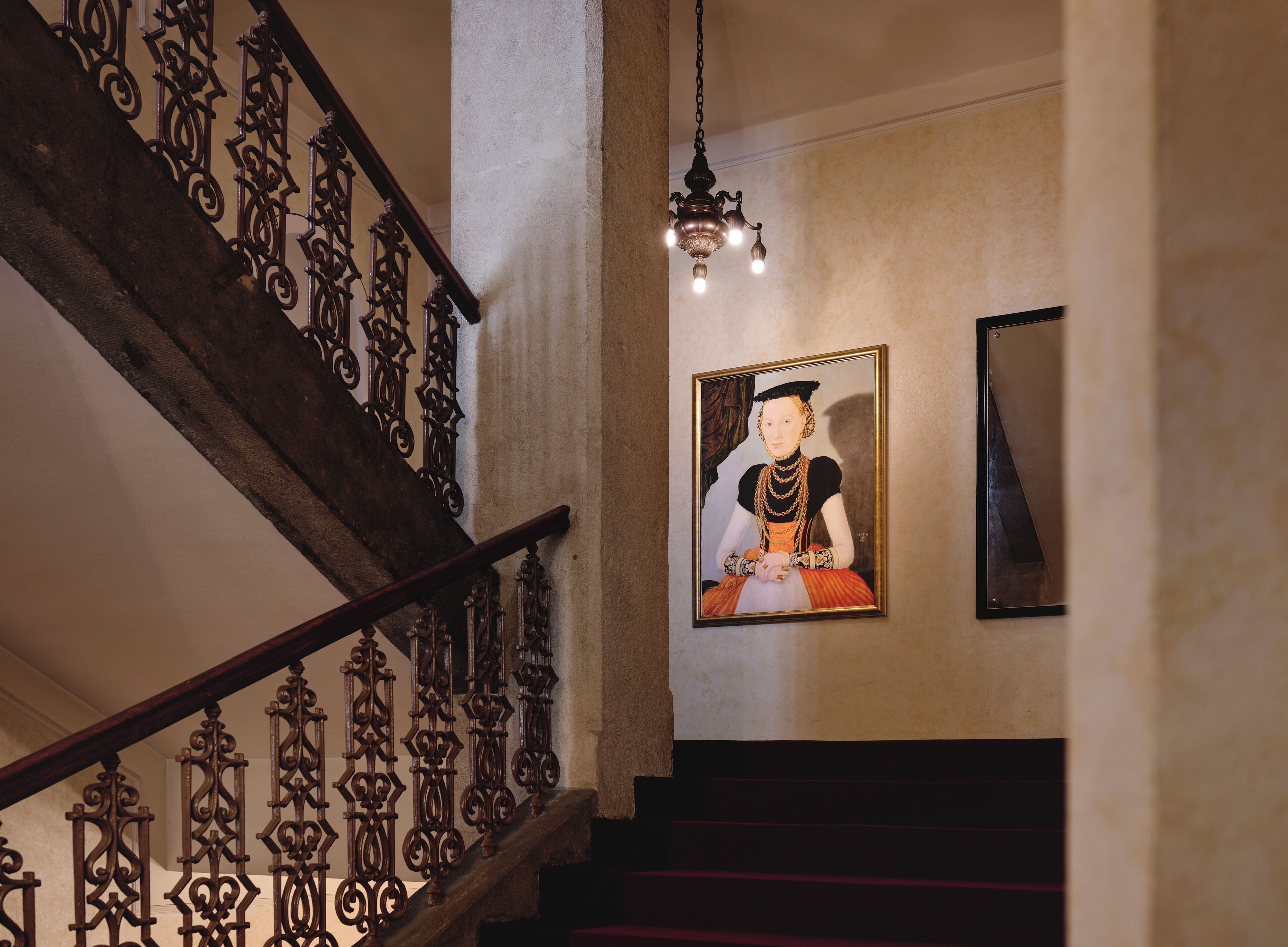 A staircase with ornate iron railings and a chandelier, a framed portrait on the wall, and a mirror.