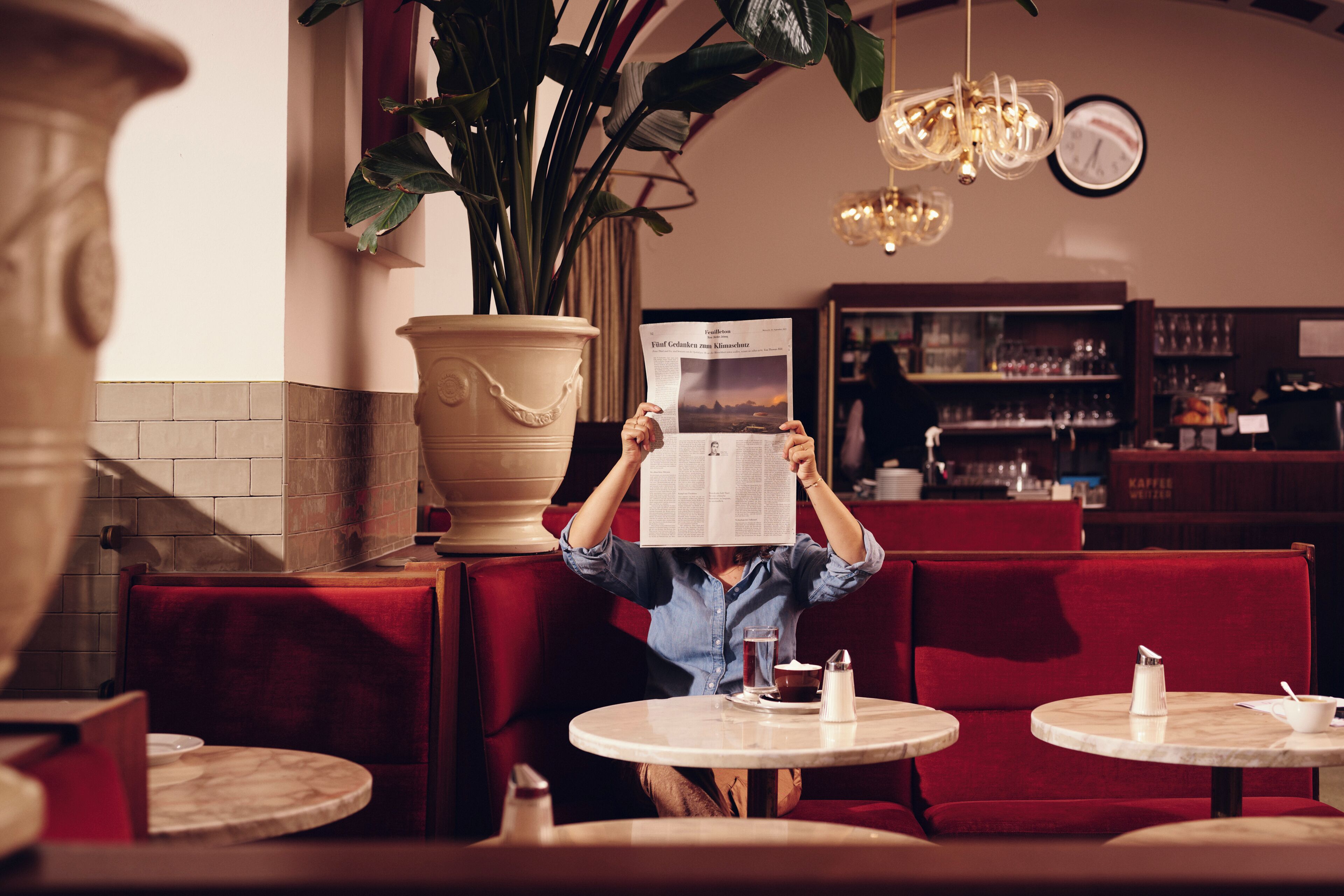 A person reading a menu in a restaurant with red seating, a large potted plant, and a clock on the wall.