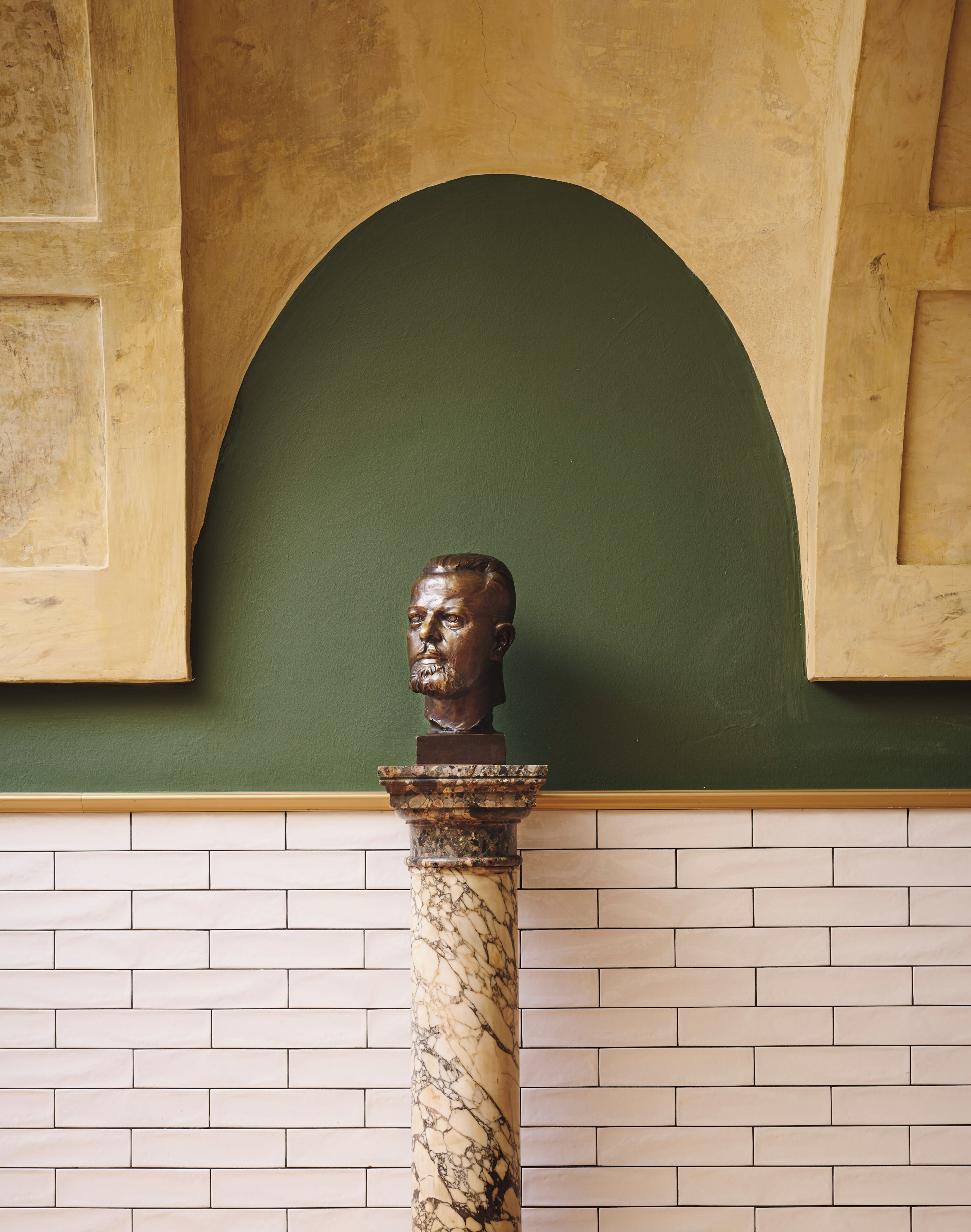 Busto sobre un pedestal de mármol frente a una pared verde con base de ladrillo.