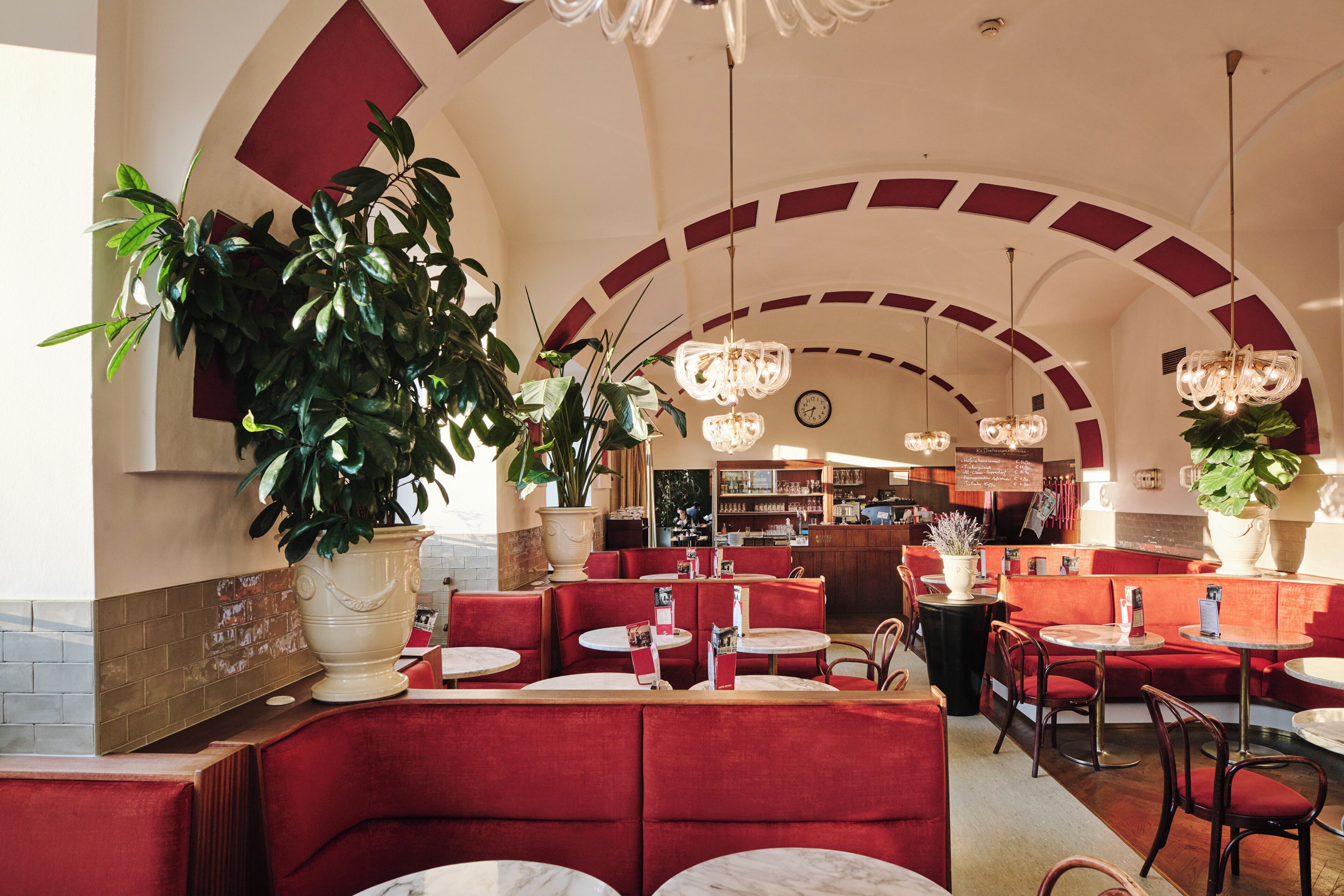 Ein Restaurantinneres mit roten Sitzgelegenheiten, weißen Tischen und großen Kübelpflanzen.