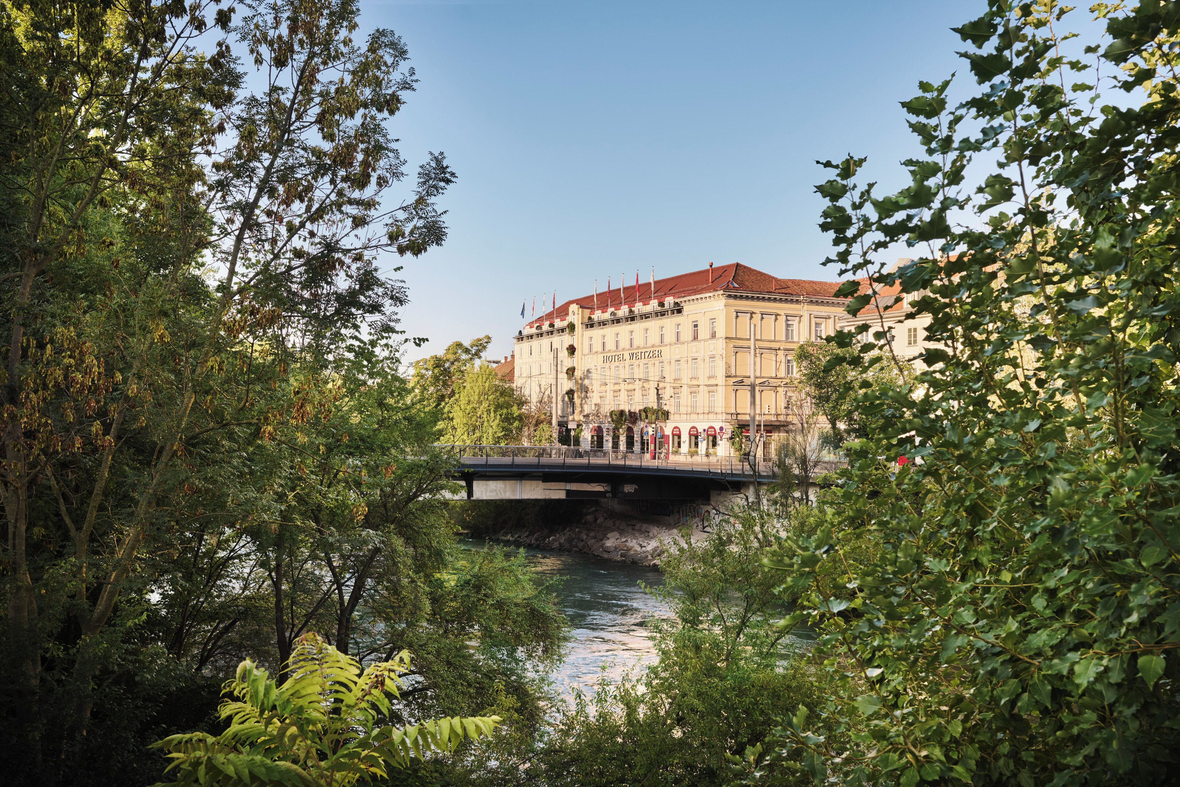 A hotel with a red roof and classical architecture, a bridge over a river, and lush greenery.