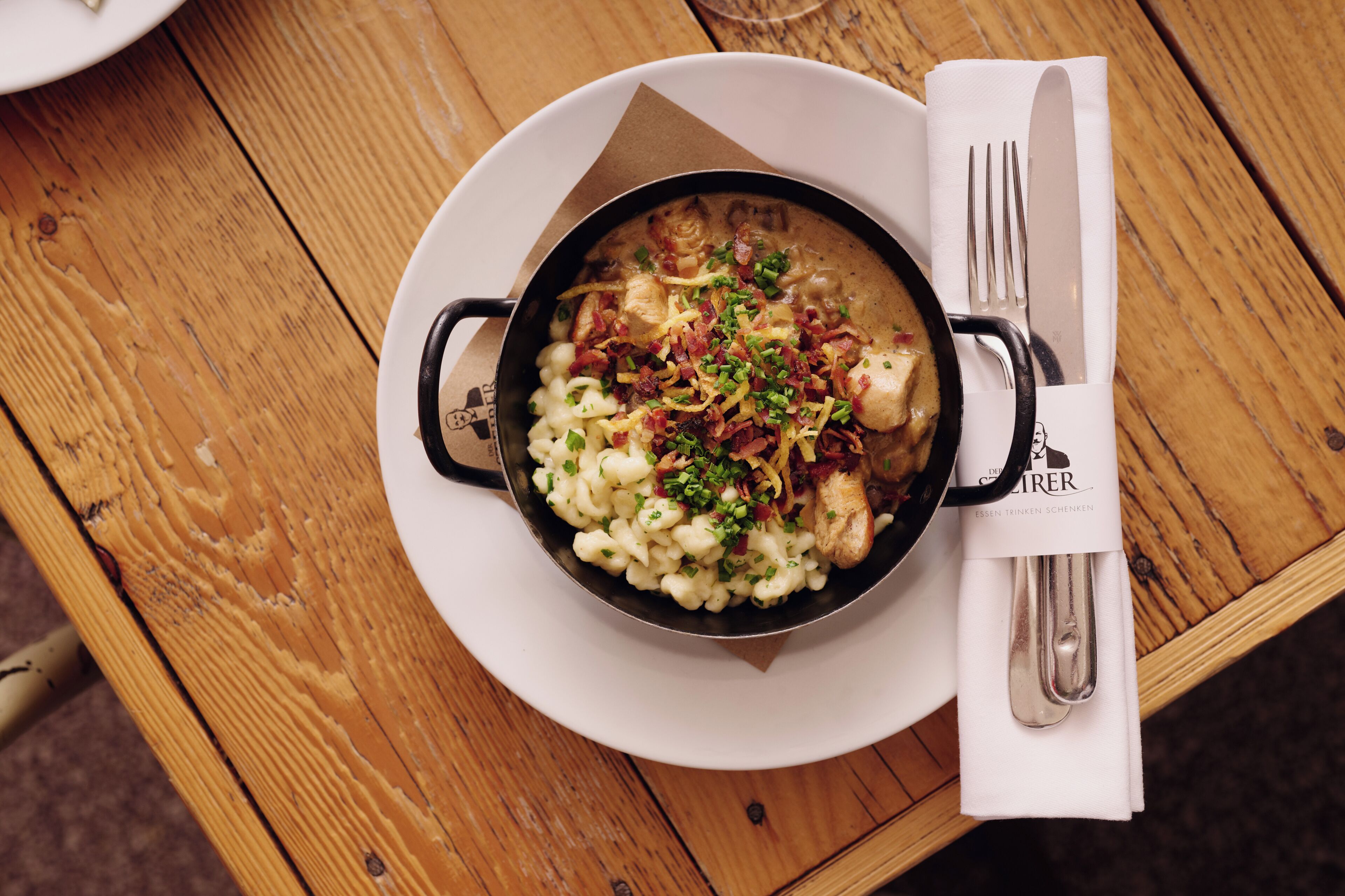 Eine Schüssel mit Essen auf einem weißen Teller, dazu ein Servietttuch und Besteck.