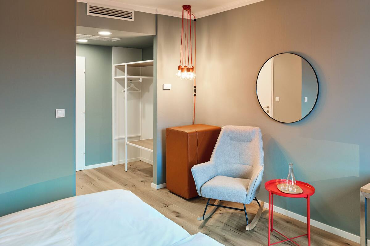 A hotel room with a bed, a wardrobe, a rocking chair, a small table with glasses, and a round mirror on the wall.