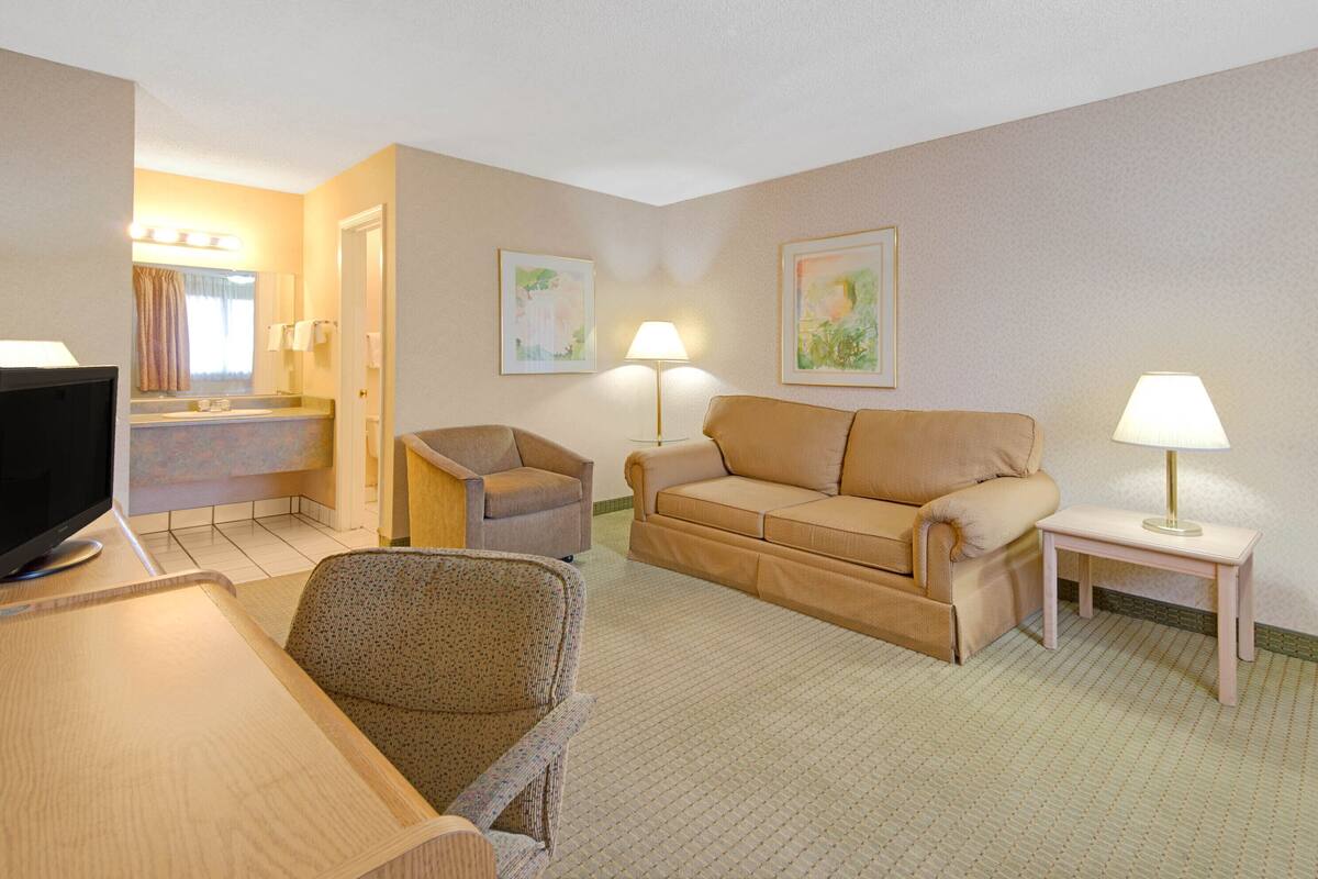 A hotel room with a sofa, armchair, a small table, a TV, and a lamp.