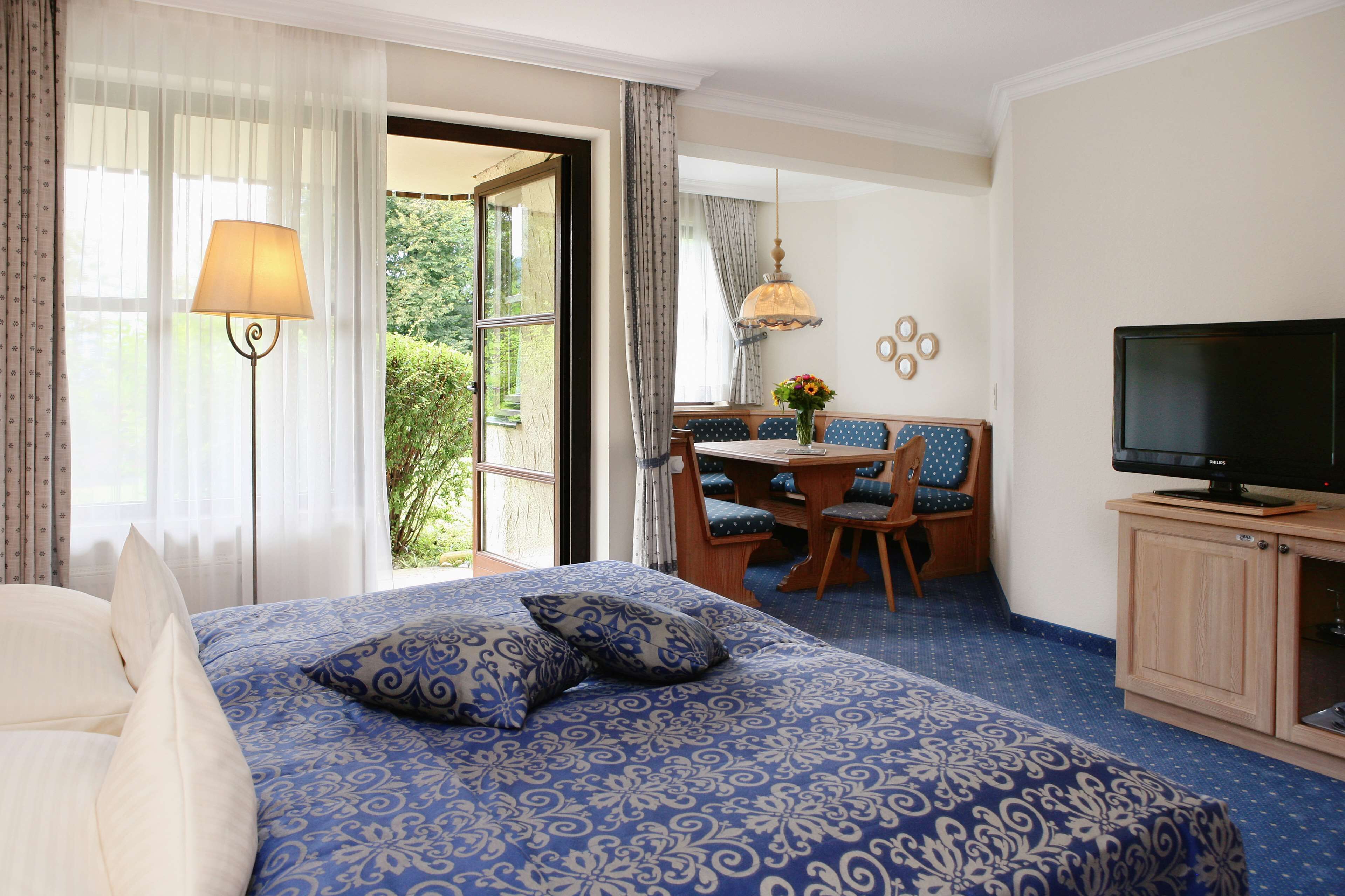 A hotel room with a large bed, a desk with a chair, a television, and a view of greenery outside.