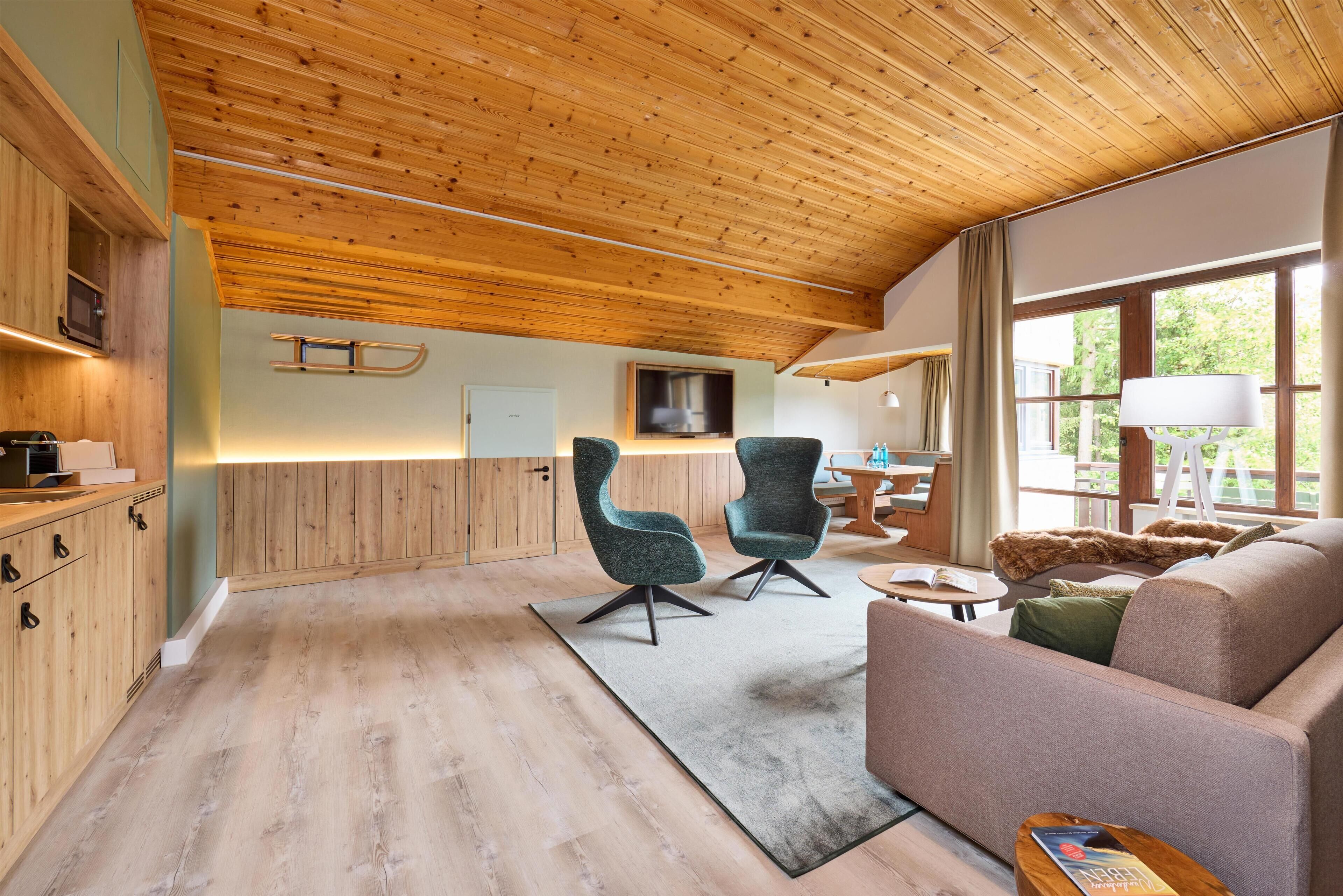 Un interior moderno con vigas de madera en el techo, un sofá, dos sillones, una mesita y una ventana con cortinas.