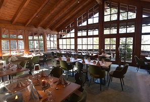 Un comedor espacioso con vigas de madera y ventanas amplias, mesas dispuestas con copas de vino y servilletas.
