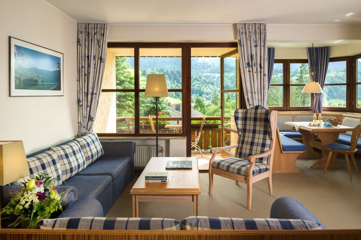 Un salon chaleureux avec un canapé, un fauteuil et une table, de grandes fenêtres avec des rideaux et une vue sur la verdure.
