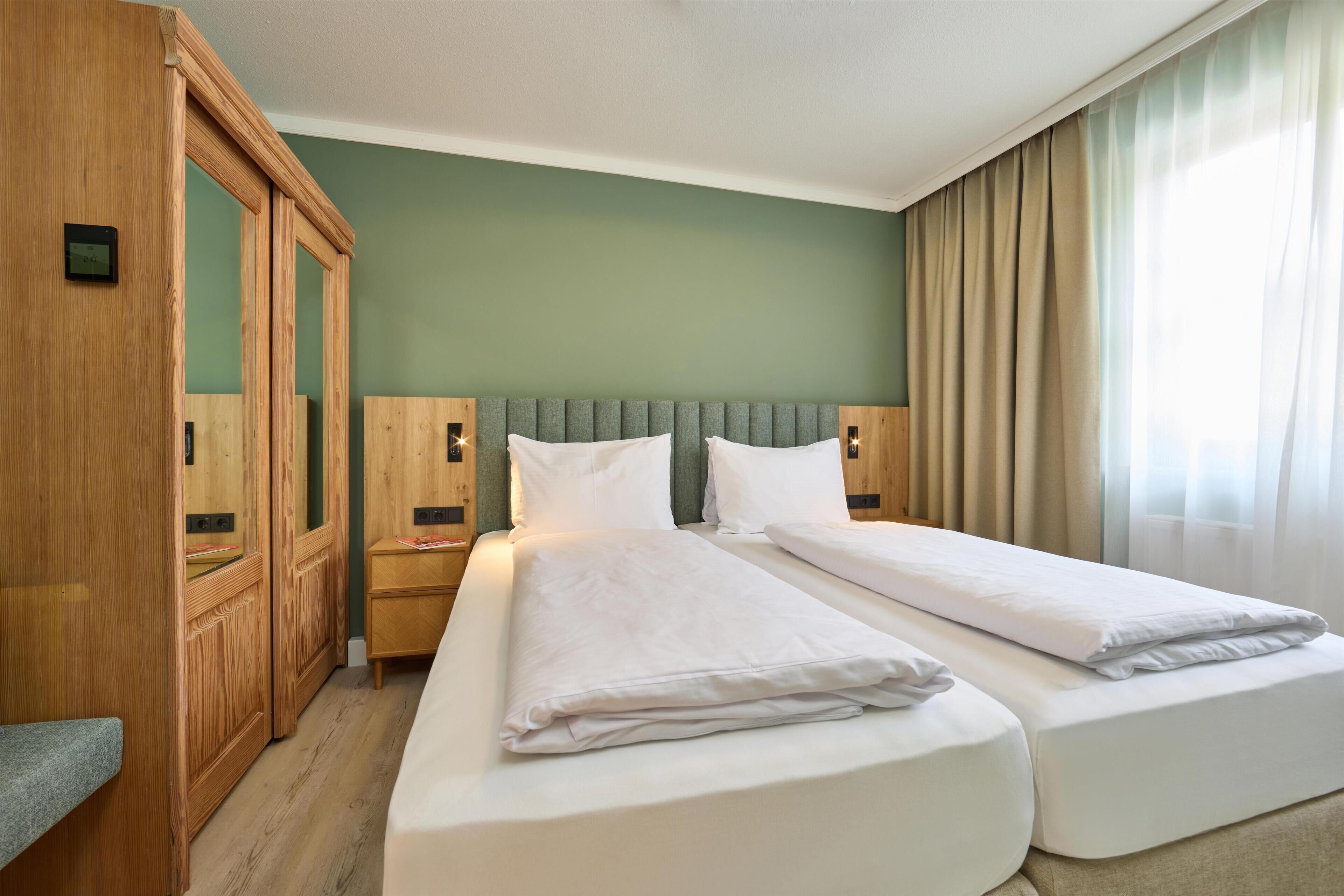 Ein Hotelzimmer mit zwei Betten, einem Holzschrank, einem Nachttisch und einem Fenster mit Vorhängen.