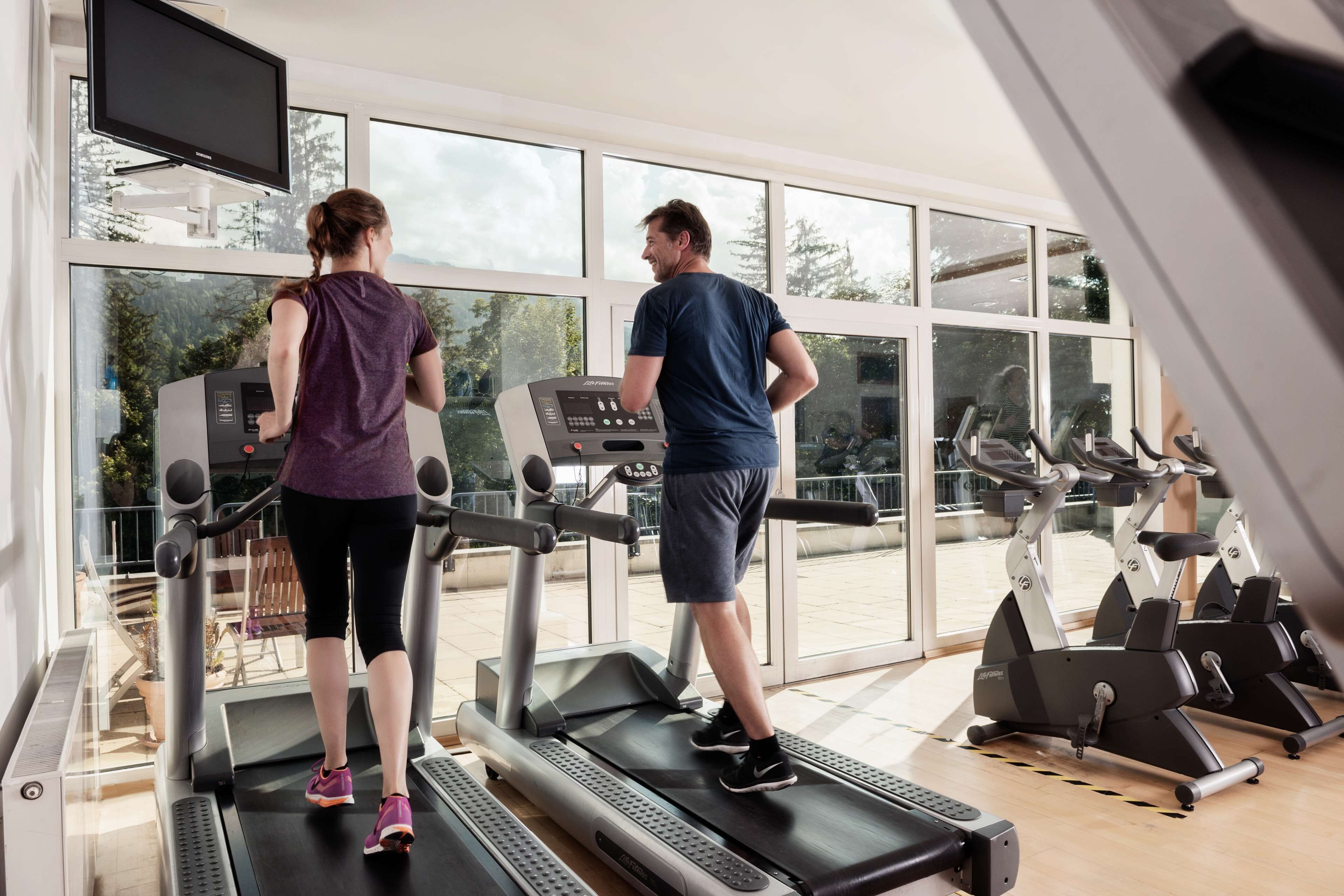Two people exercising on treadmills in a gym with large windows and a television mounted on the wall.