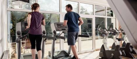Two people exercising on treadmills in a gym with large windows and a television mounted on the wall.