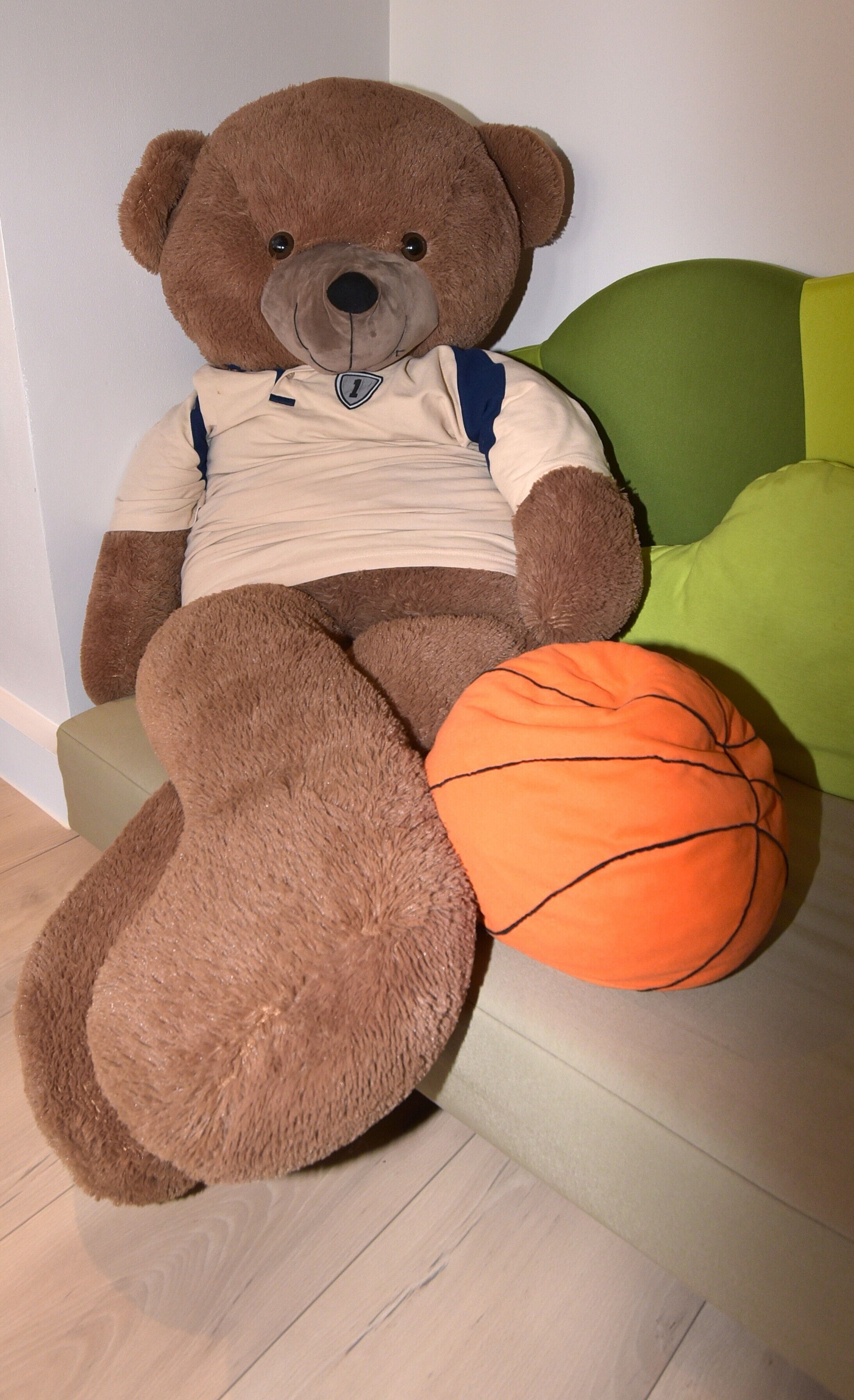 A large teddy bear sitting on a green and white couch with a basketball beside it.