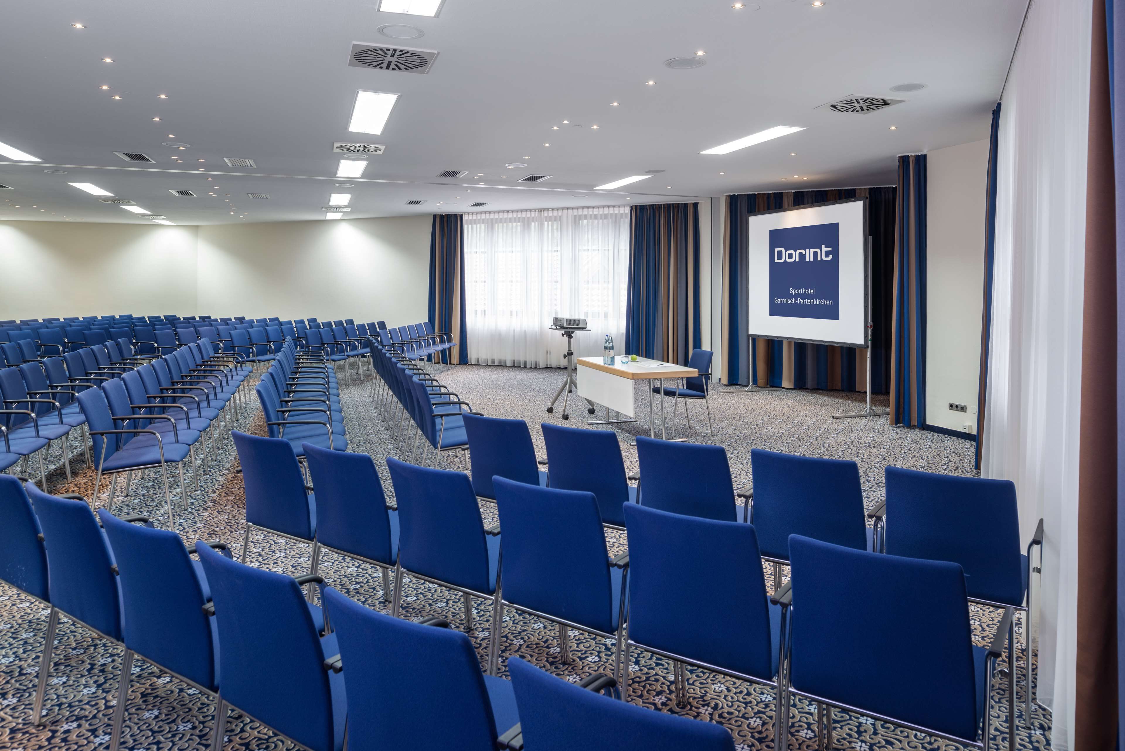 A conference room with rows of blue chairs, a projector screen, and a presentation area.