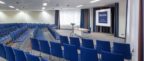 A conference room with rows of blue chairs, a projector screen, and a presentation area.