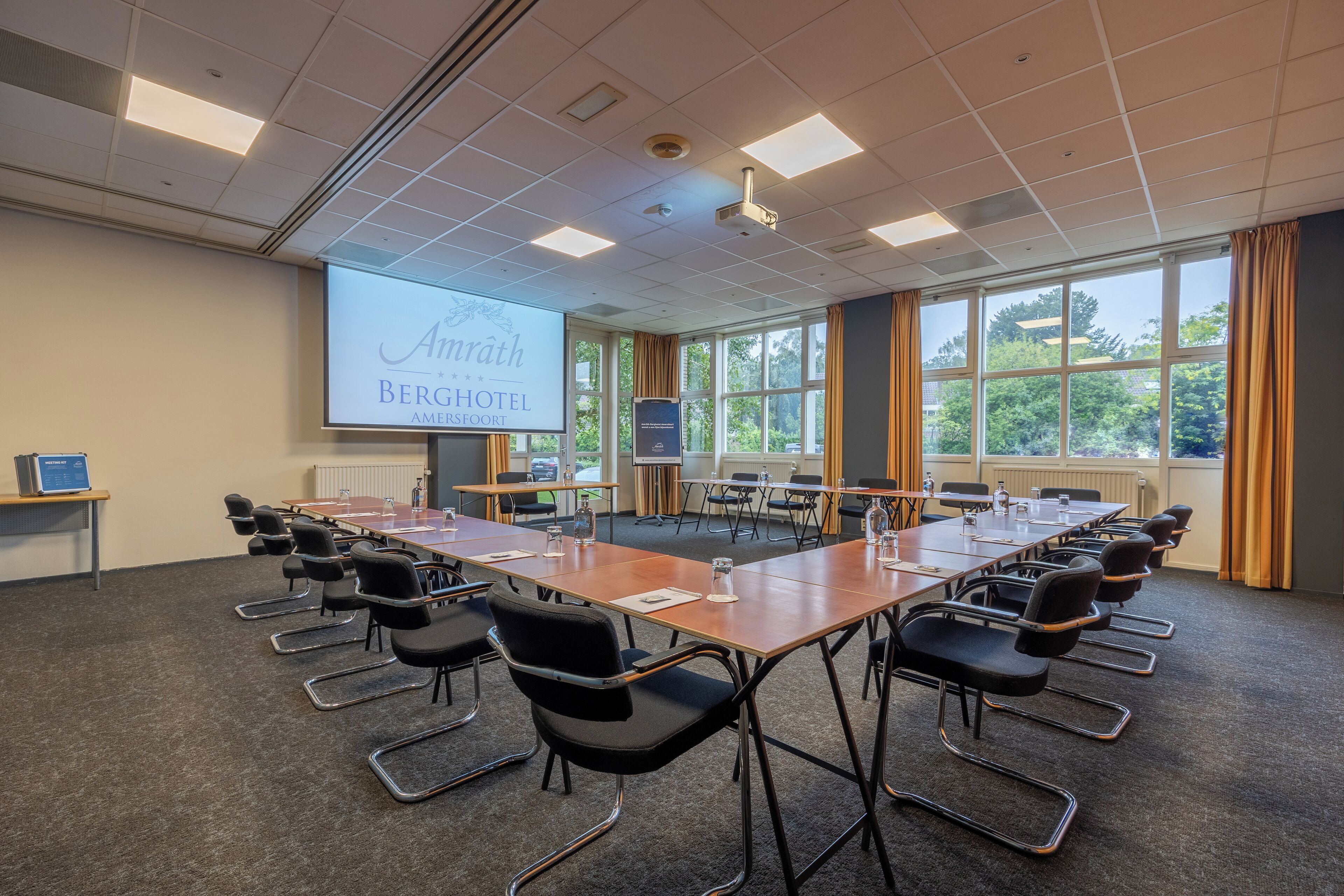 A conference room with a long table, chairs, a projector screen, and large windows with curtains.